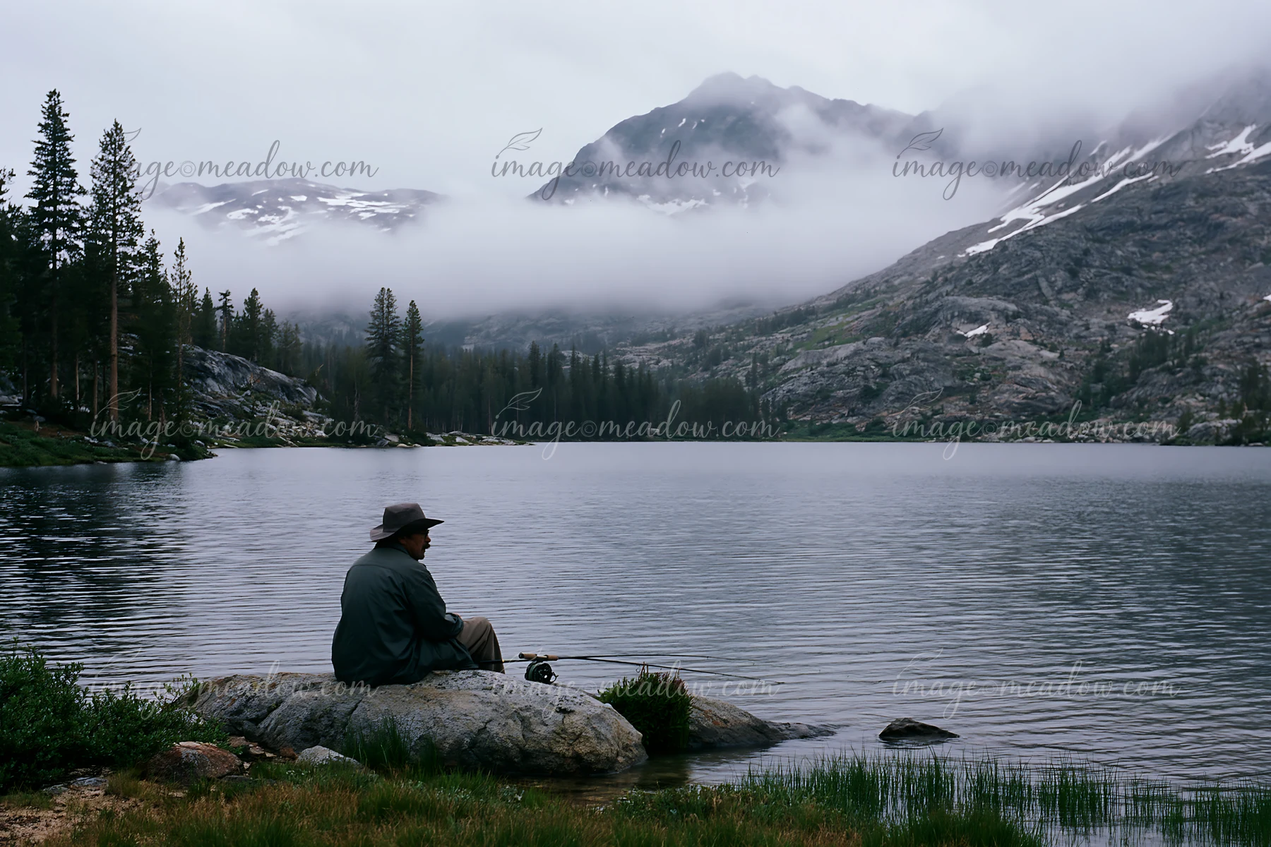 A person sits on a rock, fishing at an alpine lake beneath cloud-covered peaks.