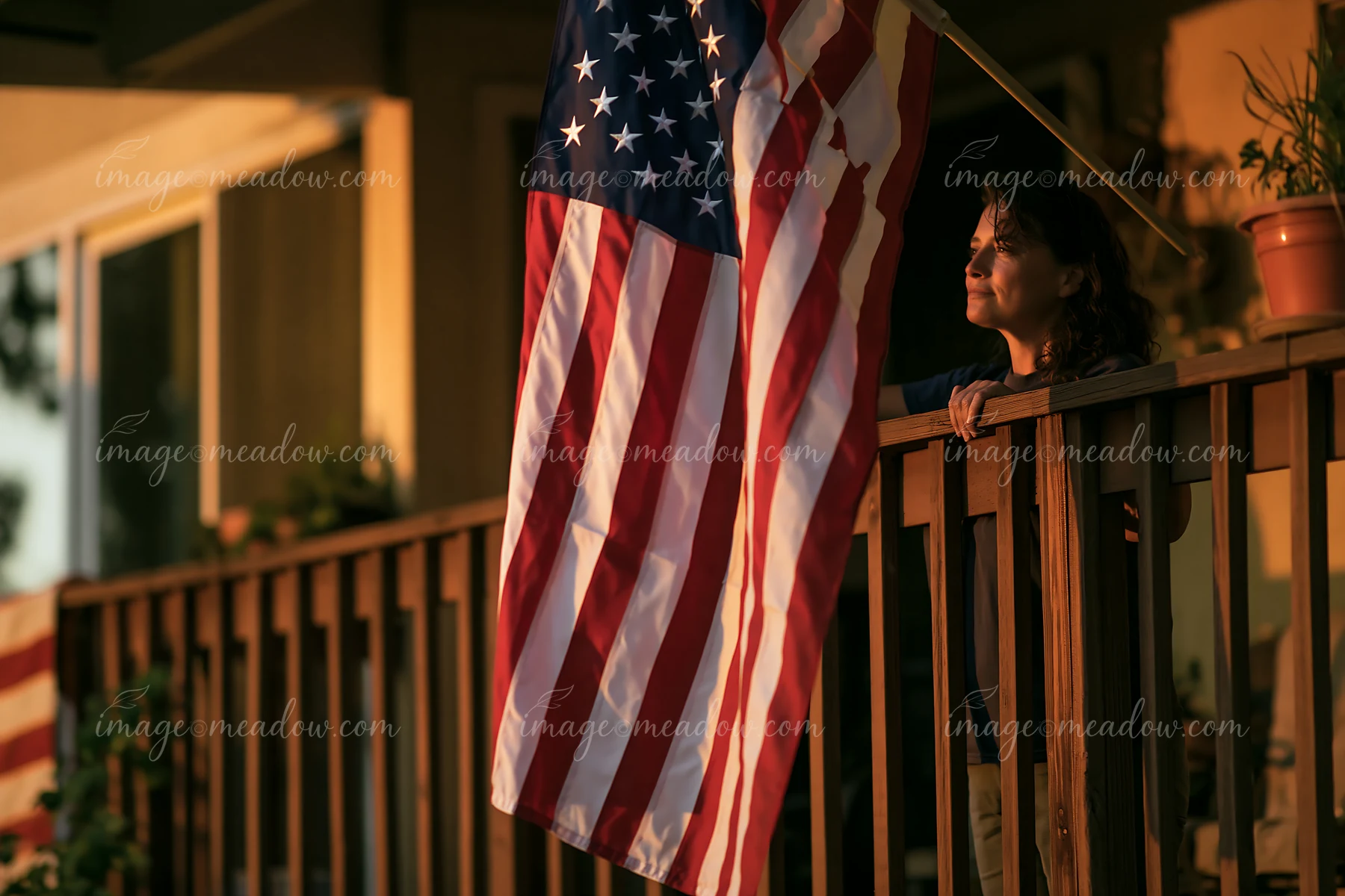 A person stands on a balcony of an apartment building, looking out with an American flag hanging beside them.