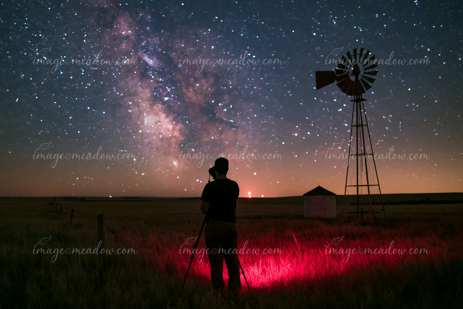 A person using camera under Milky Way stands on a grassy field beside a windmill.
