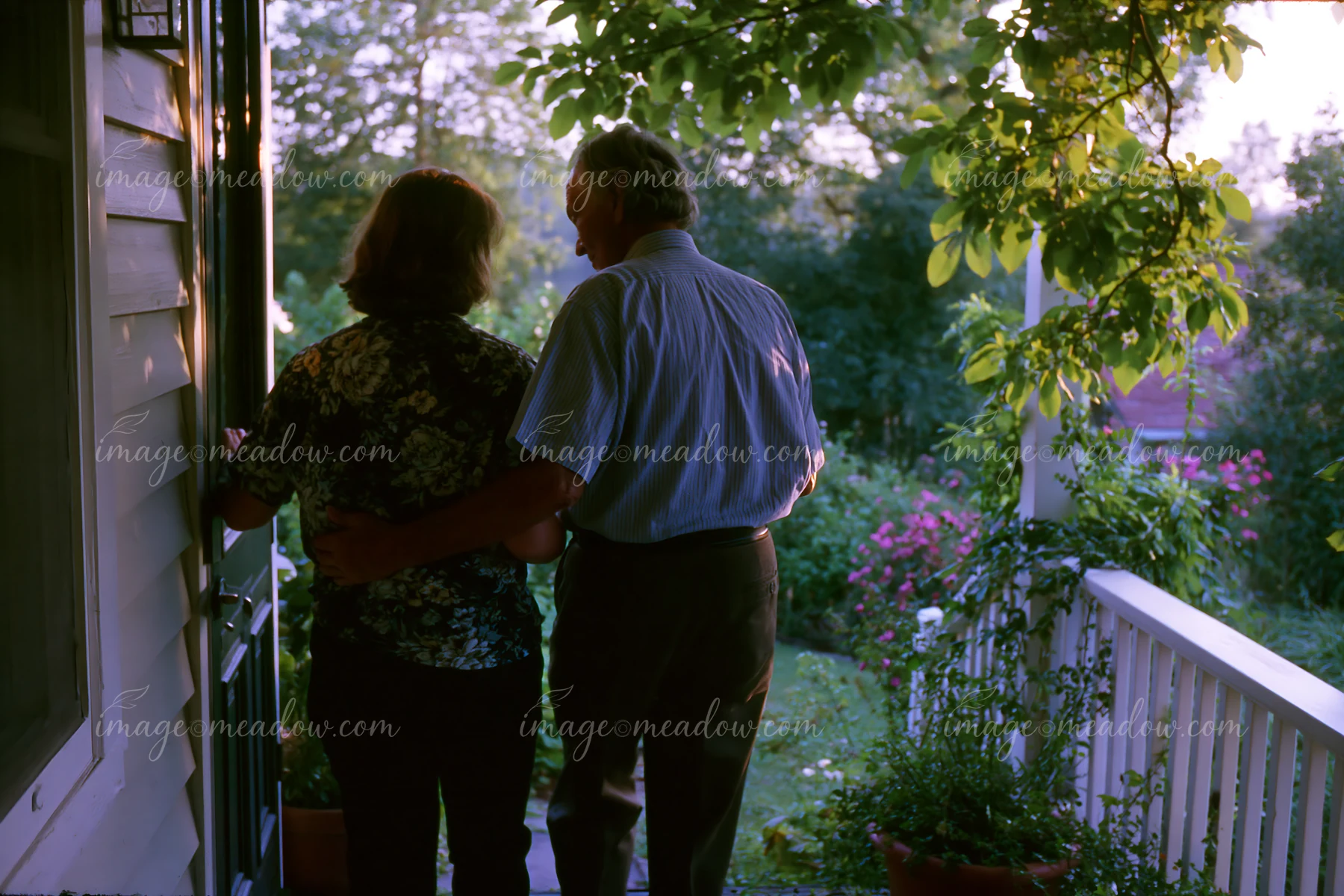 A person with Parkinson's disease receiving support stands beside another individual on a porch in a garden.