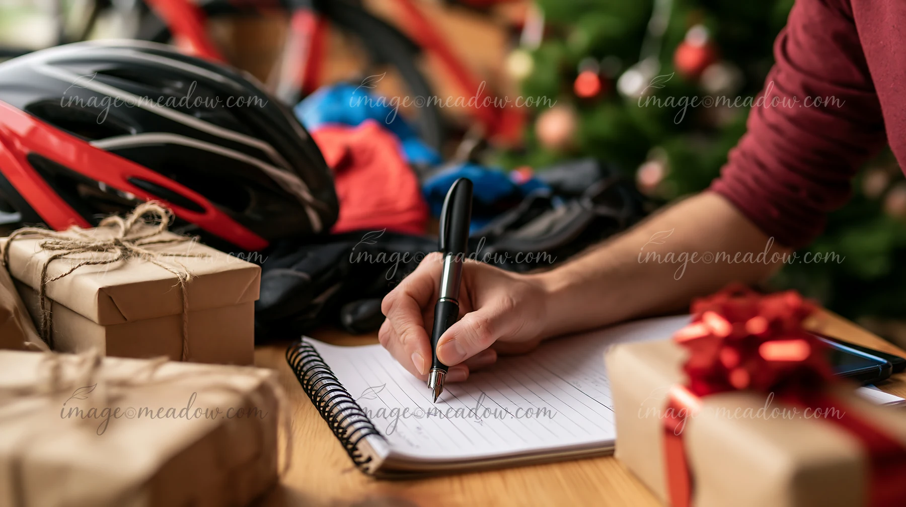 A person writes a gift list on a notebook while sitting at a desk filled with wrapped presents and a bicycle helmet.