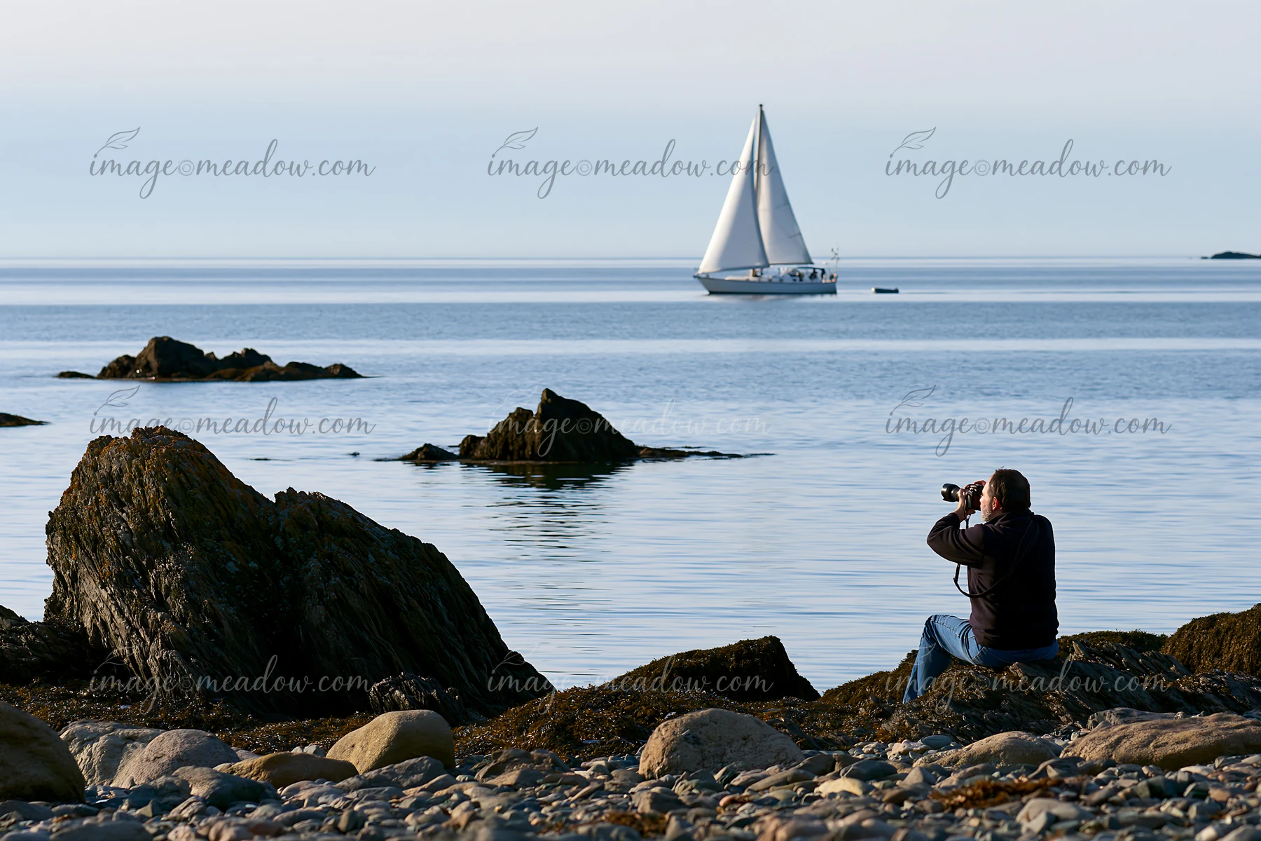 A photographer photographing a sailboat at sea from a rocky shore.