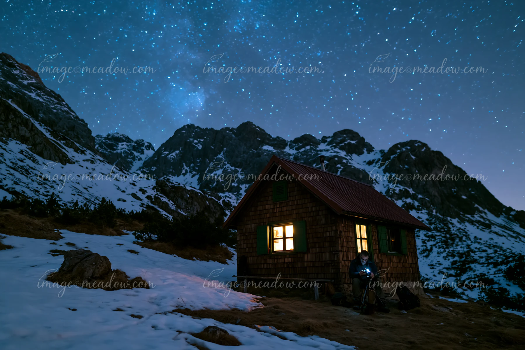 A photographer under stars in the mountains sets up a camera outside a rustic cabin.