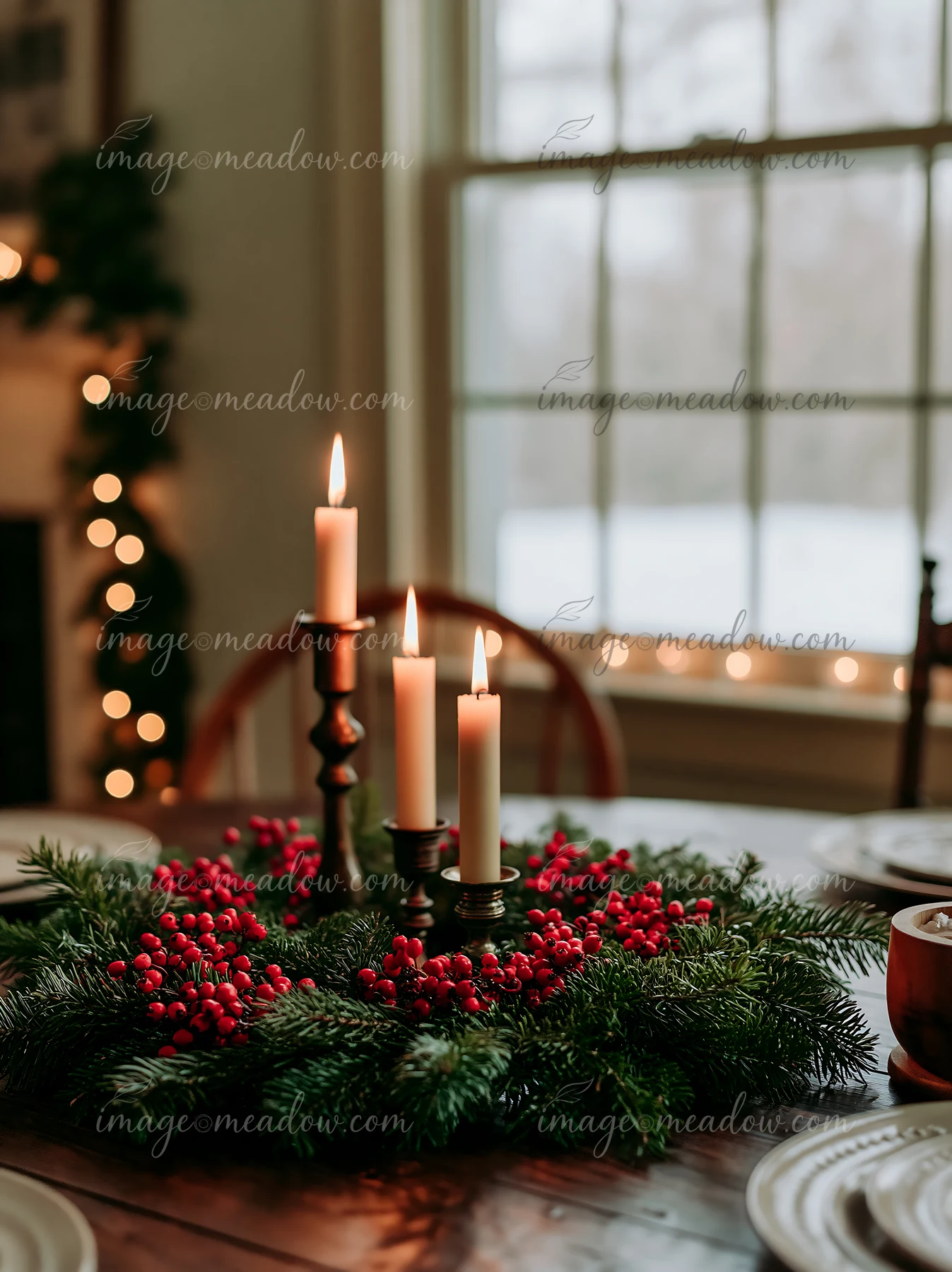 A pine wreath with red berries sits on a wooden table with three burning candles.