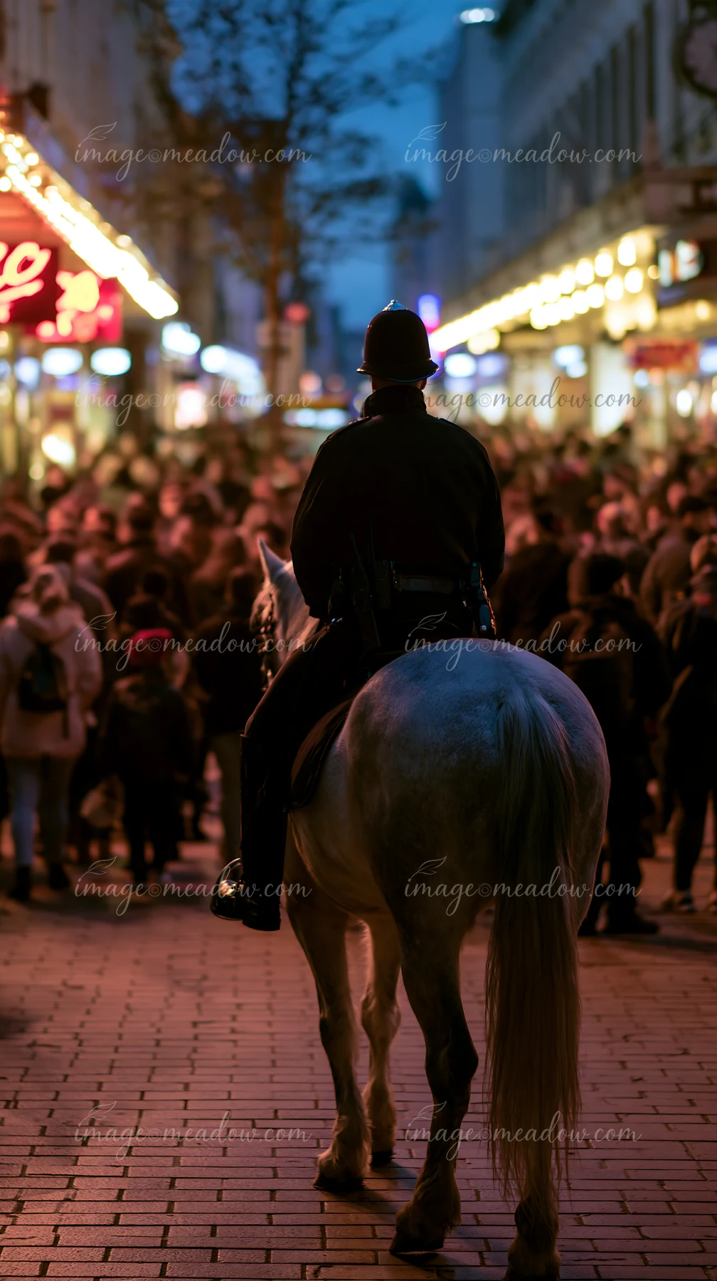 A police horse walks beside a crowded pedestrian street filled with people at night.