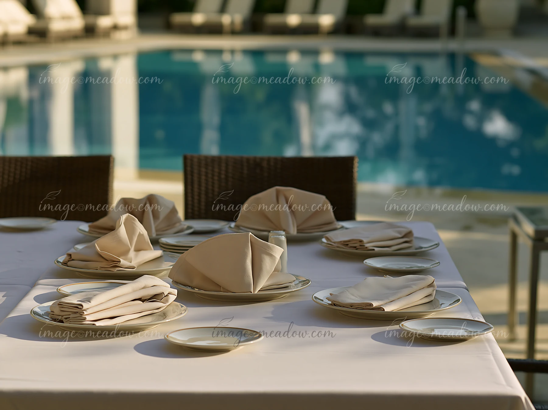 A poolside buffet table is arranged with plates and folded napkins beside a clear blue swimming pool.