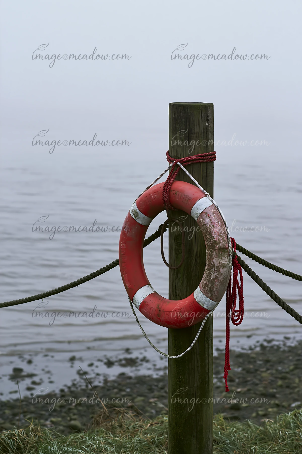 A red and white lifebuoy hangs on a wooden post along a foggy coastline.
