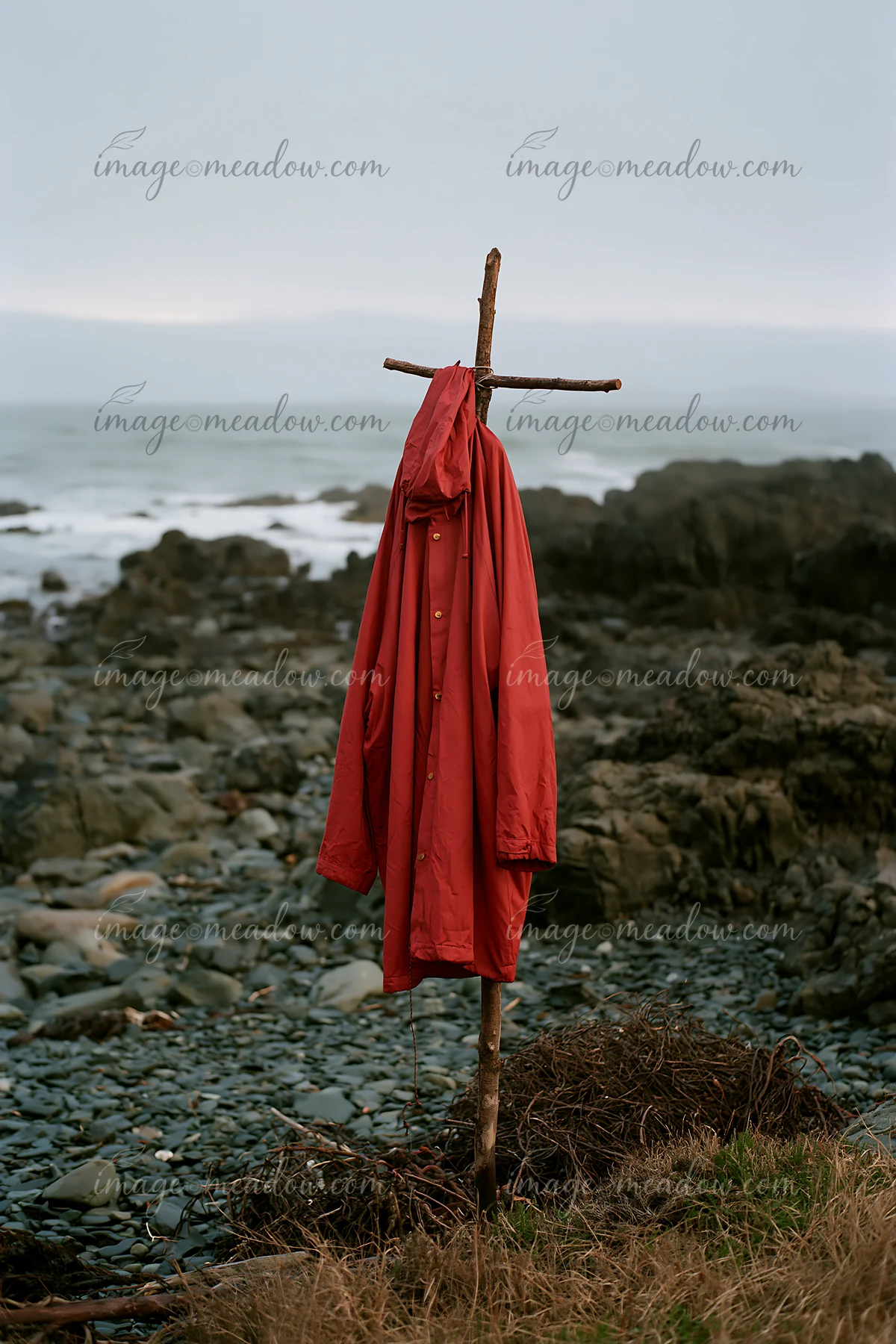 A red coat is draped over a stick, positioned near the ocean at a rocky coastline with exposed rocks.