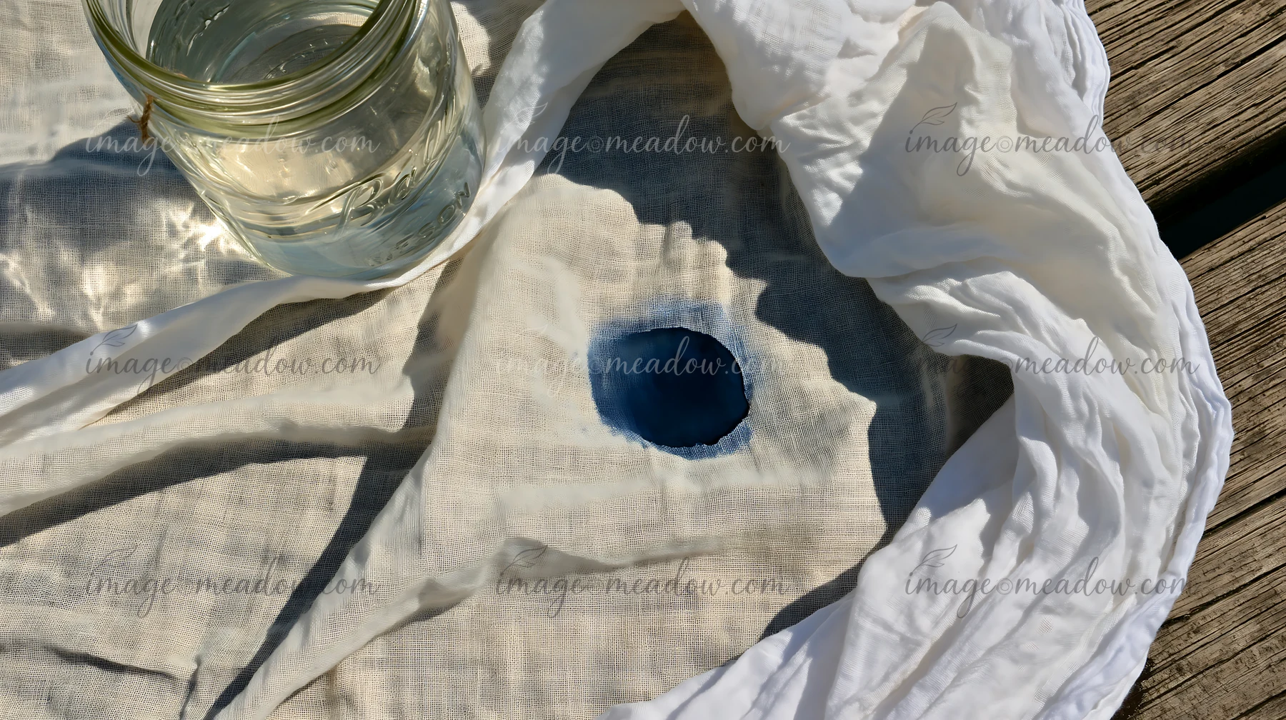 A round watercolor stain on light muslin fabric sits next to a glass jar filled with clear water.