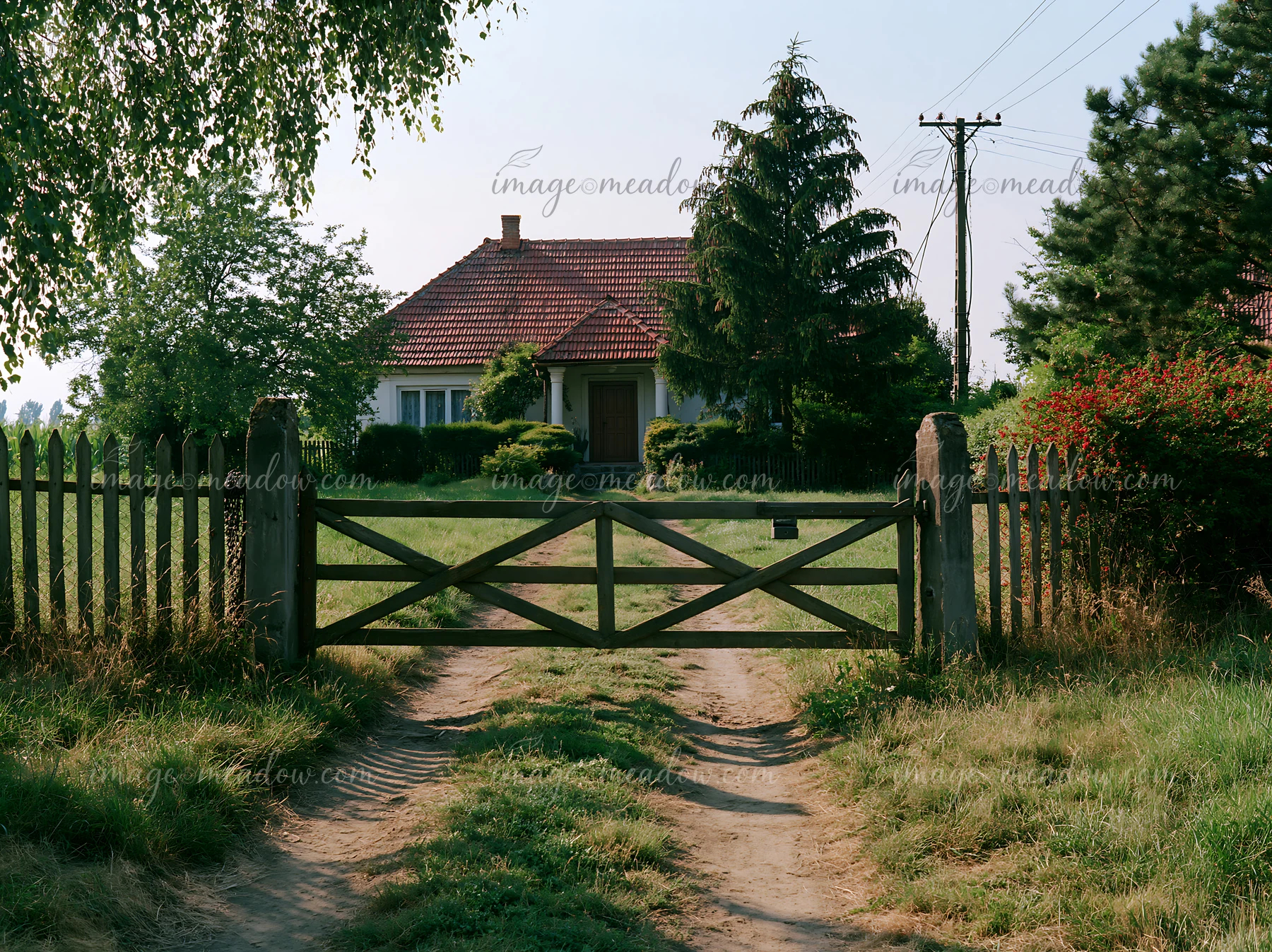 A rural cottage is visible through a wooden gate, surrounded by grass and trees.