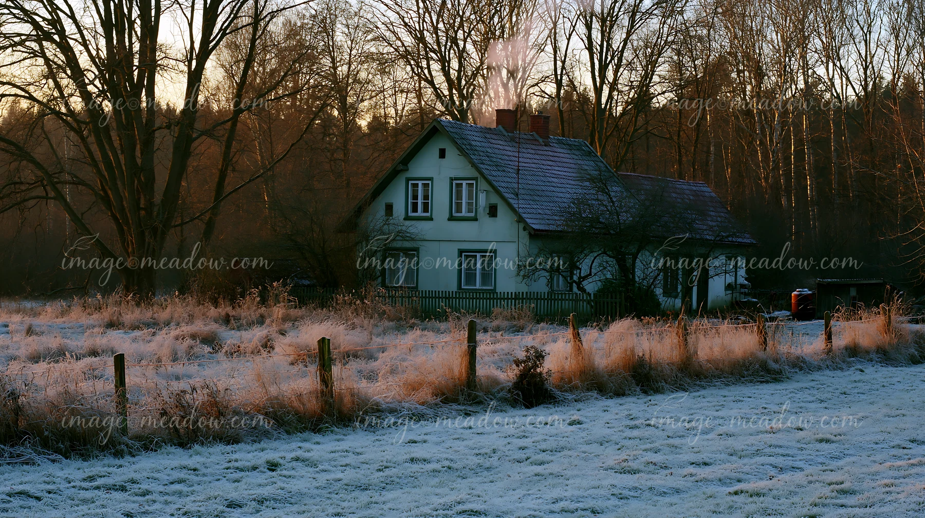 A rural cottage with chimney smoke stands in a frost-covered field surrounded by trees.