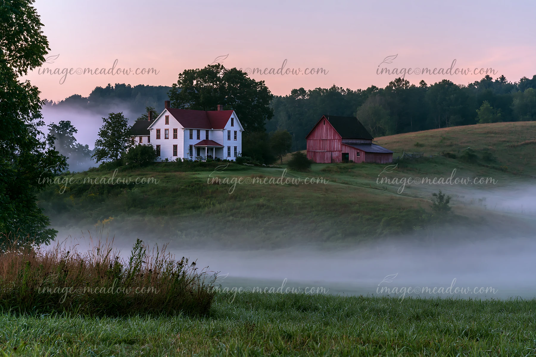 A rural farmhouse in morning fog sits on a hill, surrounded by fields and trees.