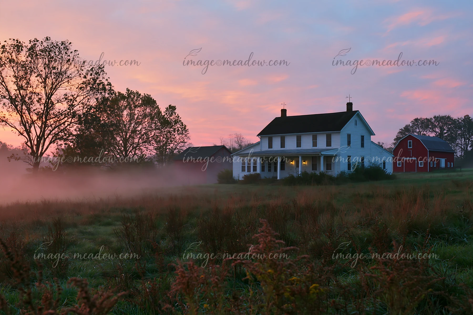 A rural farmhouse stands shrouded in morning fog with a red barn nearby.