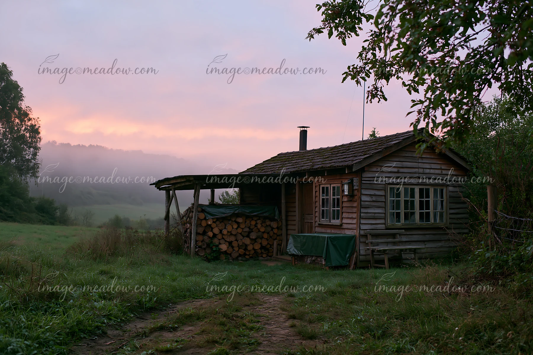 A rustic cottage stands in a meadow with a stack of firewood beside it.