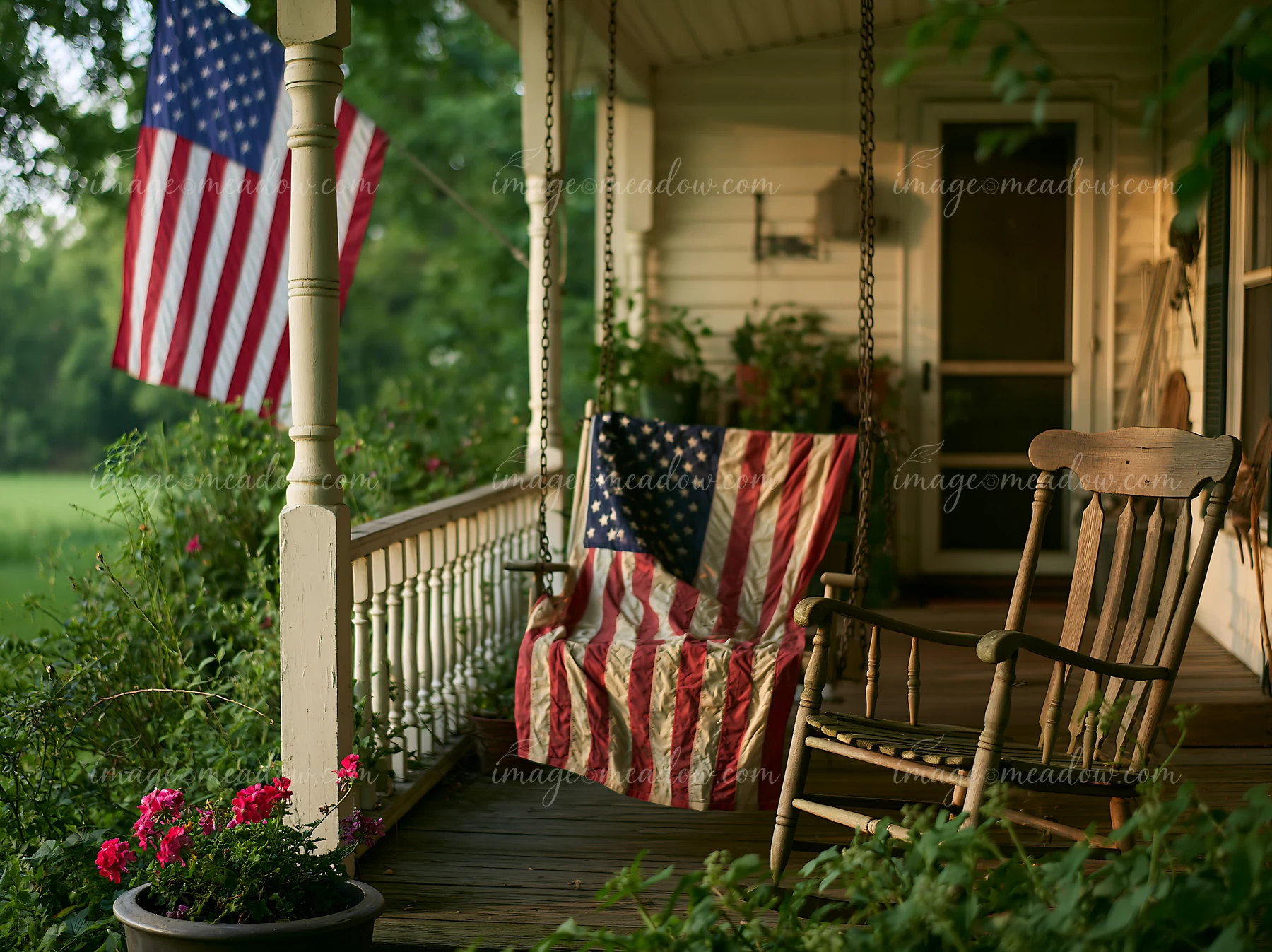 A rustic farmhouse porch displays an American flag, gently waving in the breeze alongside a wooden rocking chair and decorative potted flowers.