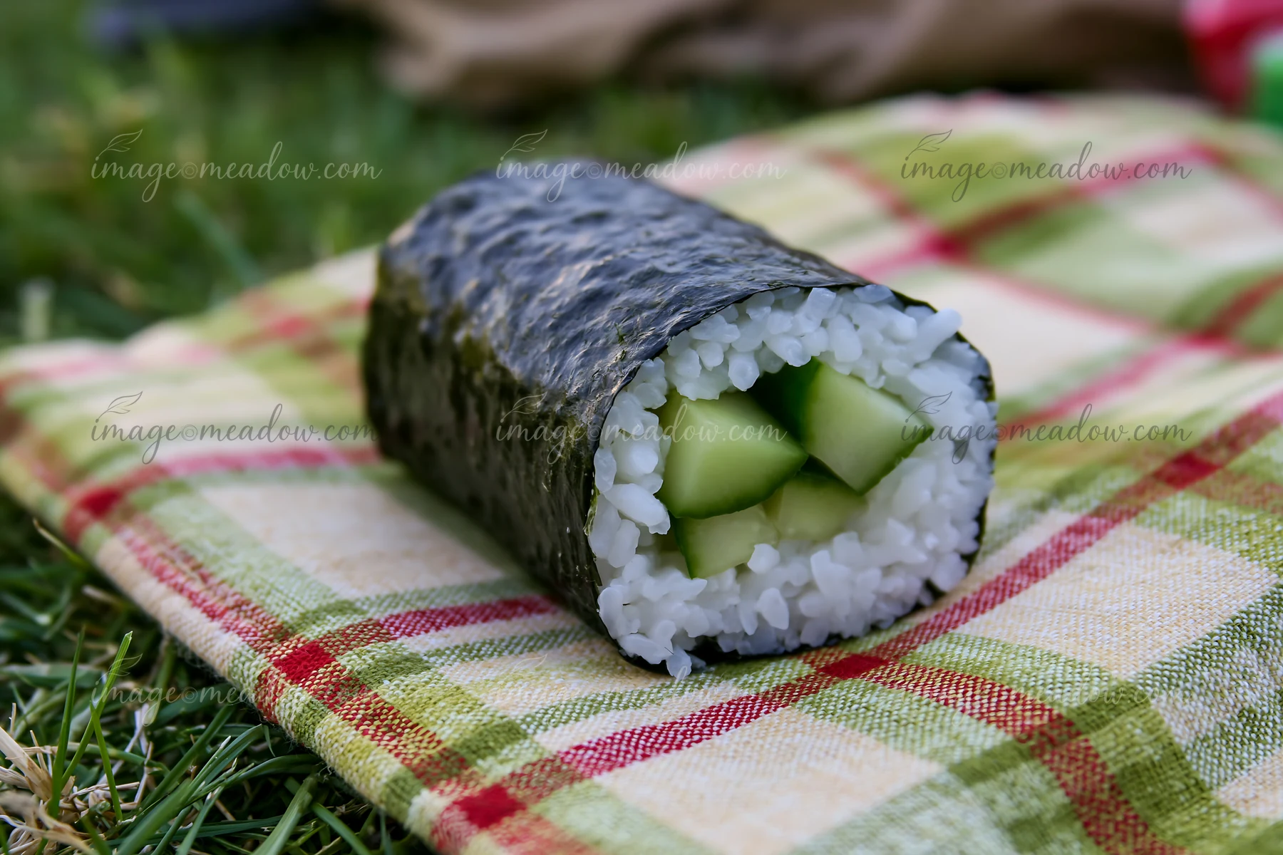 A single cucumber maki roll rests on a plaid picnic blanket, showcasing fresh cucumber pieces surrounded by sushi rice and seaweed.