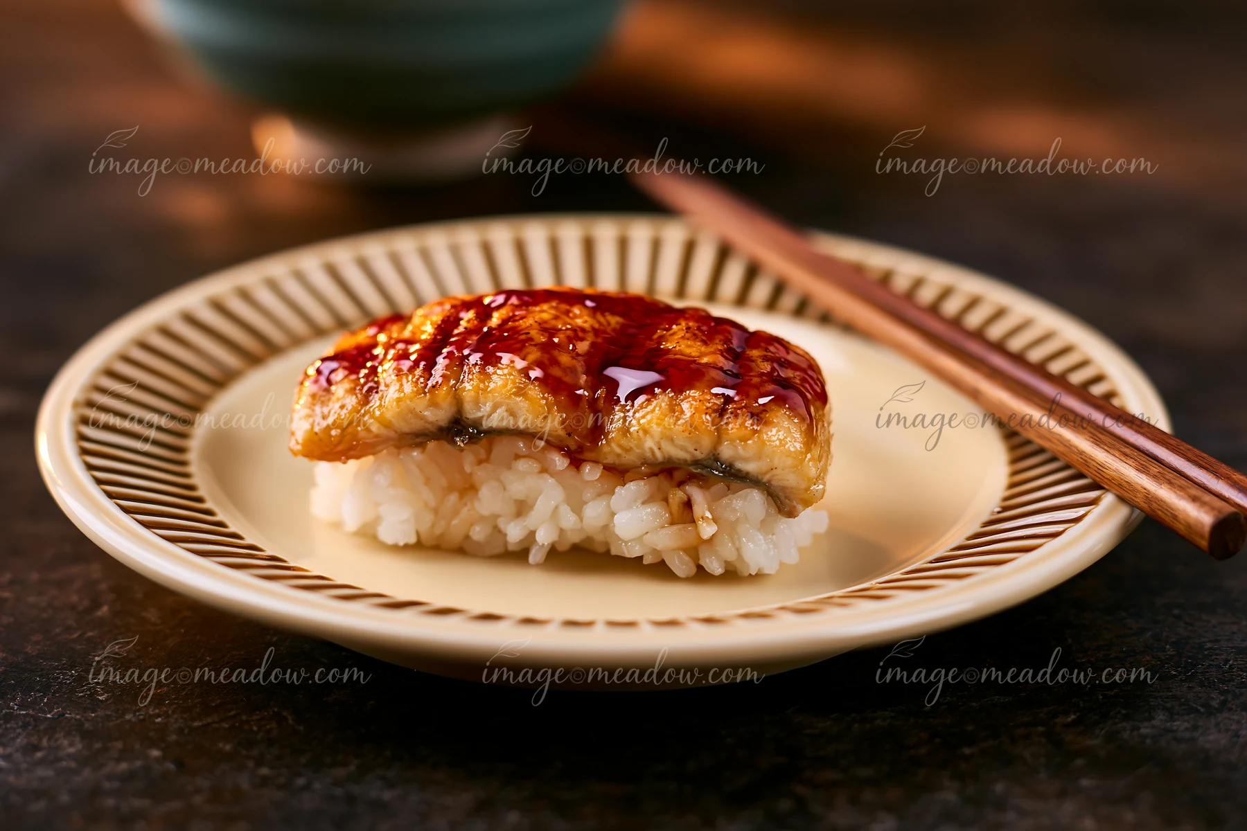 A single piece of sushi, made with grilled eel and sauce, is placed on a cream-colored plate.