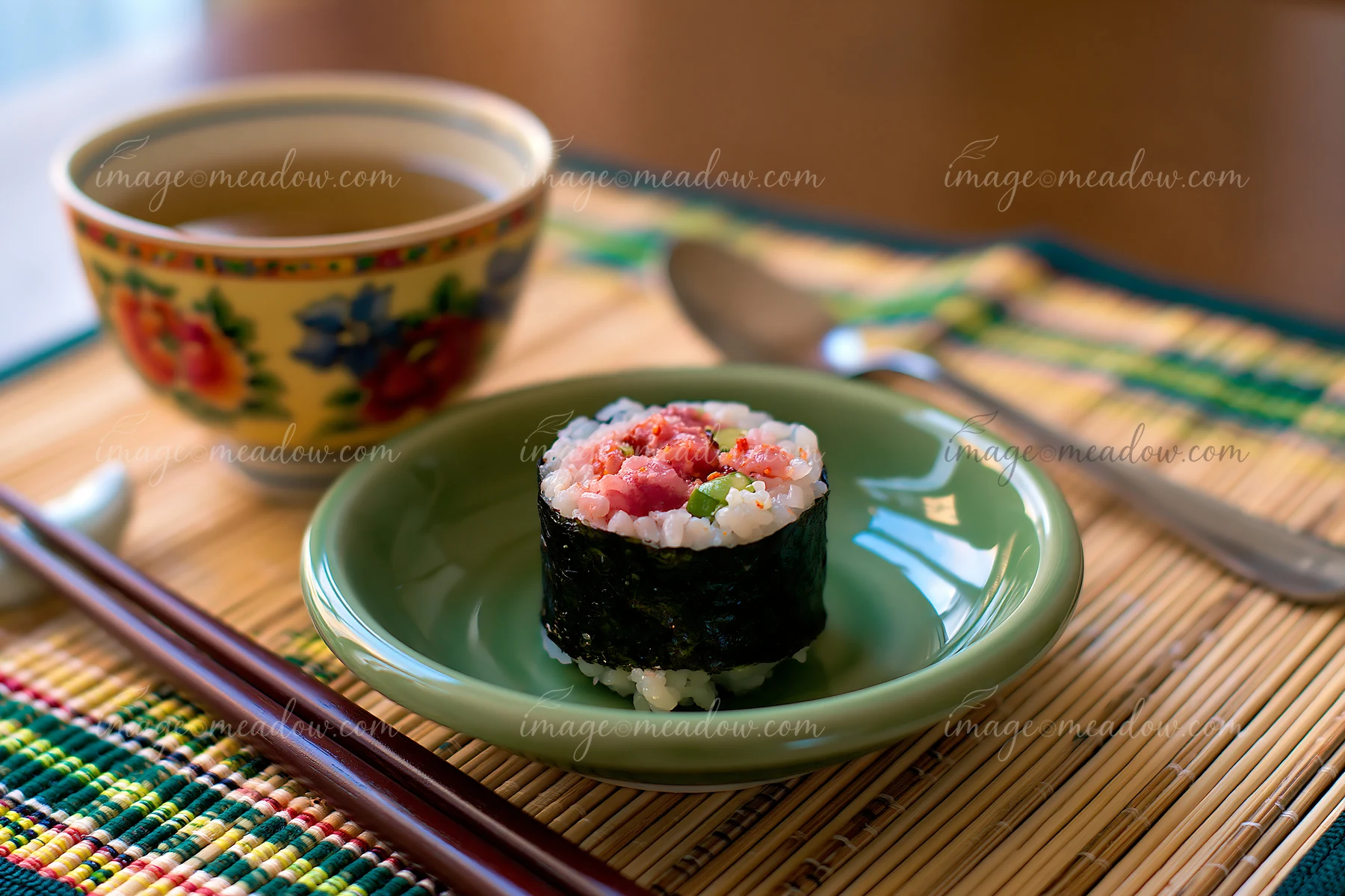 A single spicy tuna roll piece sits on a green plate next to a bowl of tea.