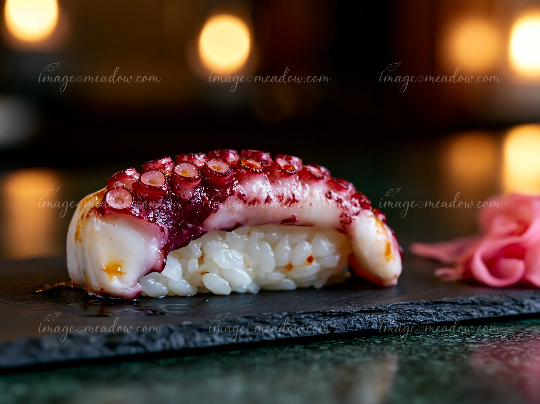 A single sushi piece on black slate features octopus on rice, served with pink pickled ginger.