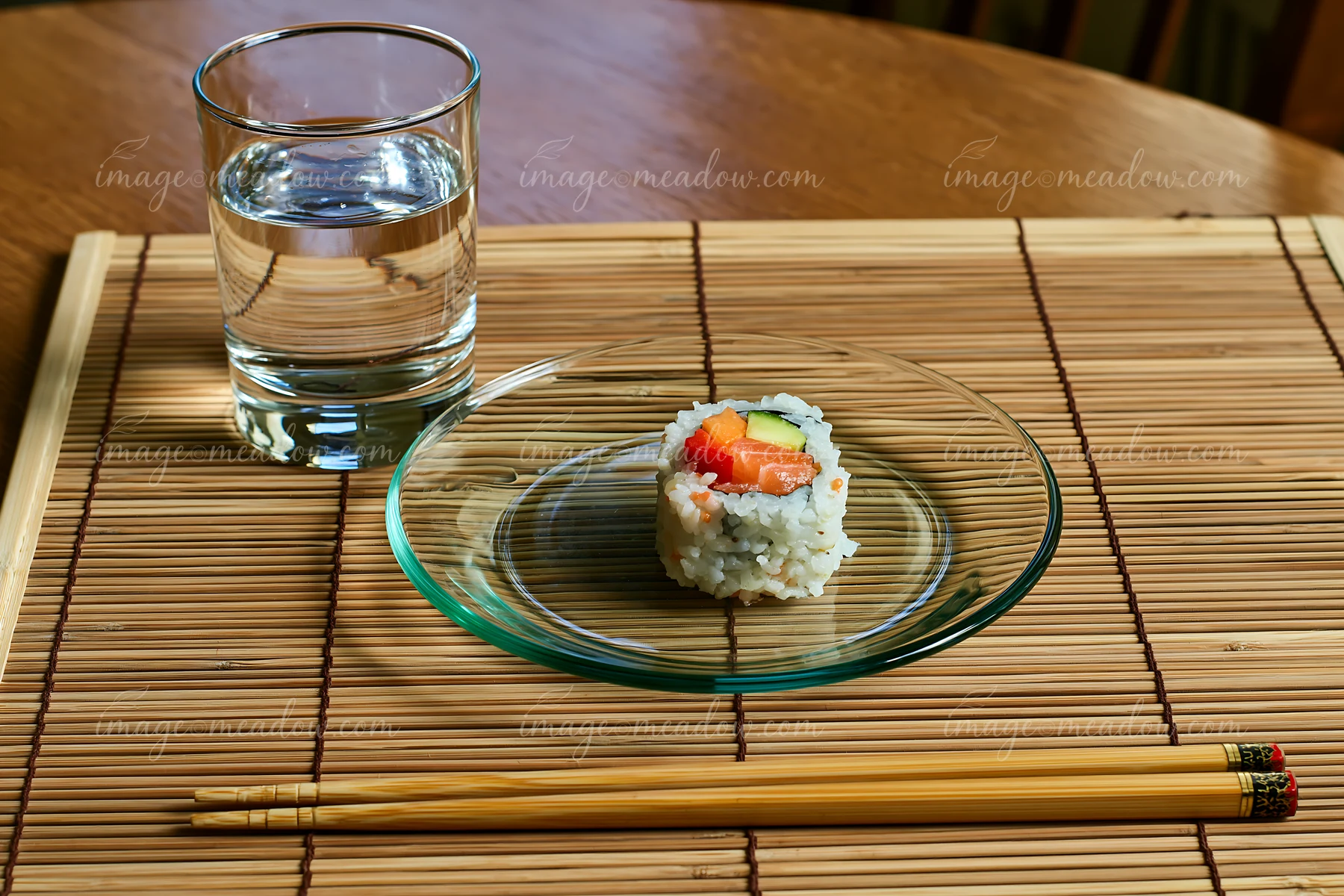 A single sushi roll on bamboo mat is placed on a clear glass plate next to a glass of water and wooden chopsticks.
