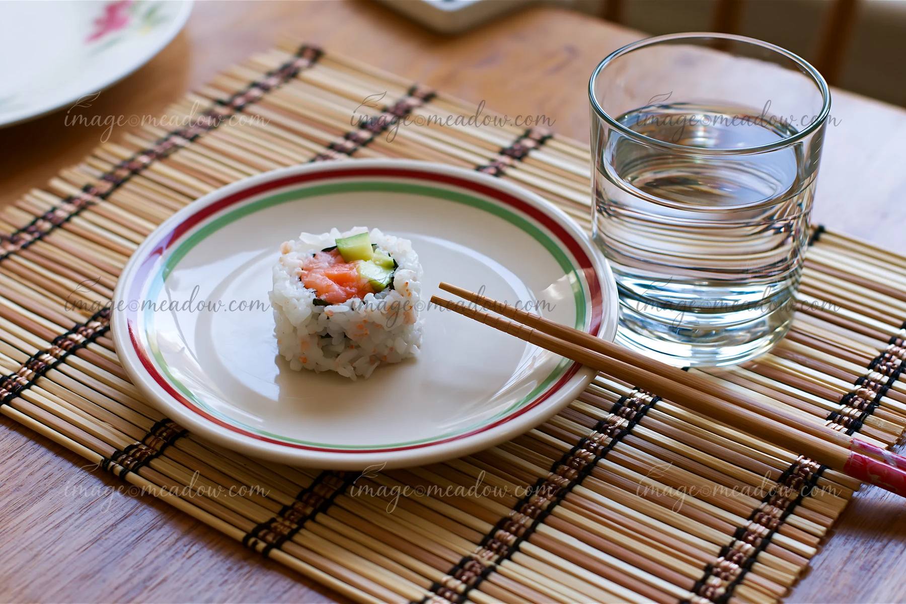 A single sushi roll on bamboo mat sits on a plate next to a glass of water and chopsticks, set on a wooden table during mealtime, showing a simple dining arrang