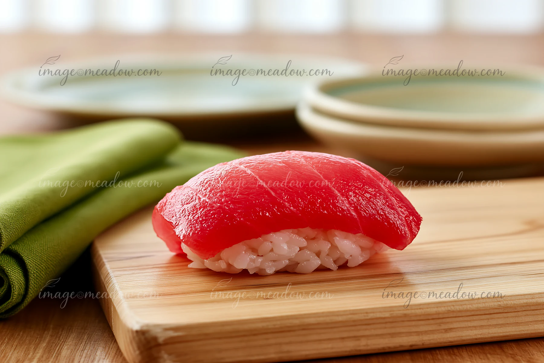 A single tuna nigiri rests on a wooden board next to green cloth and plates.
