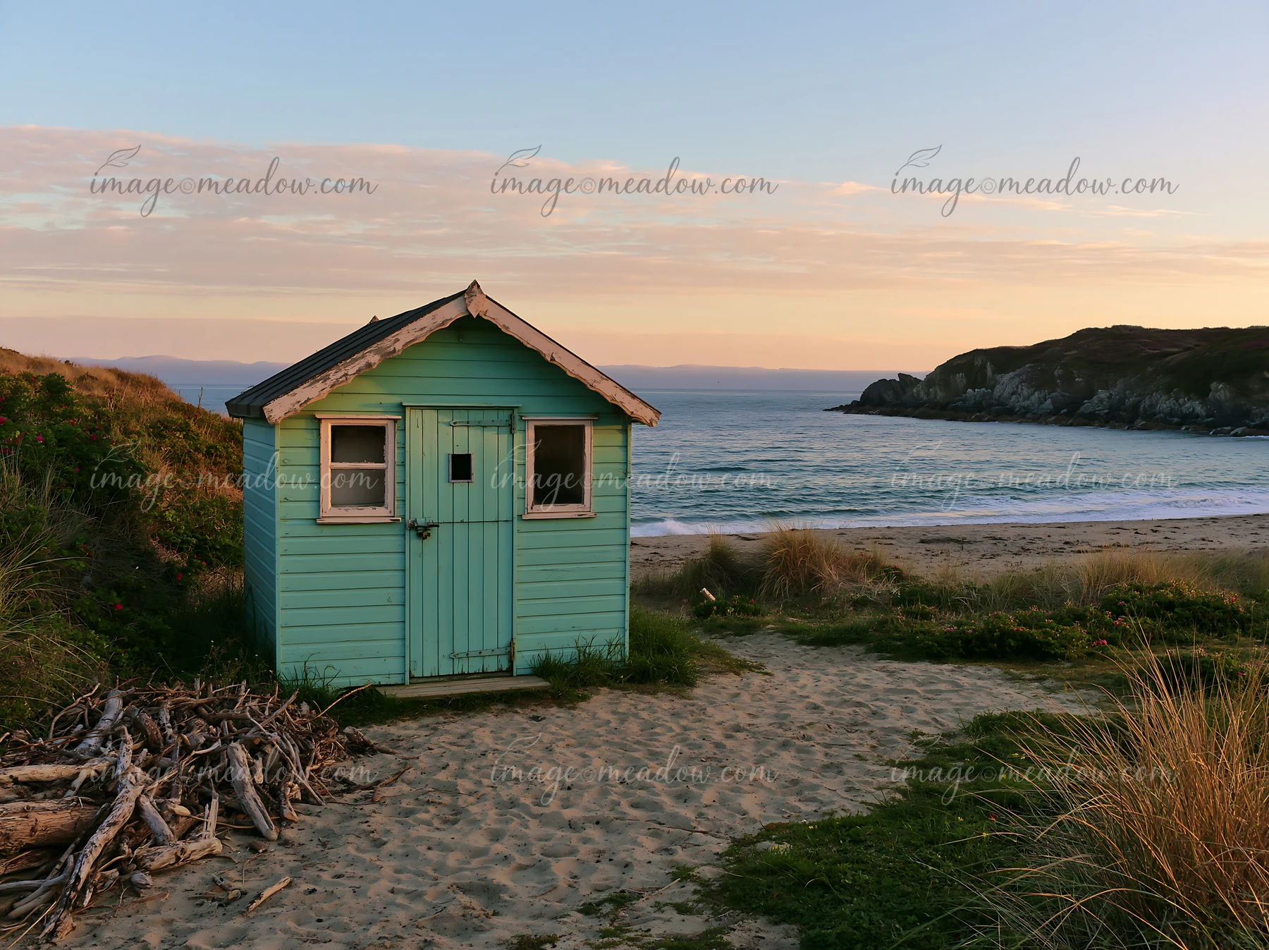 A small blue beach hut stands on the sand near a coastline with sheltered bay while waves gently lap at the shore during sunset, creating a warm glow in the sky