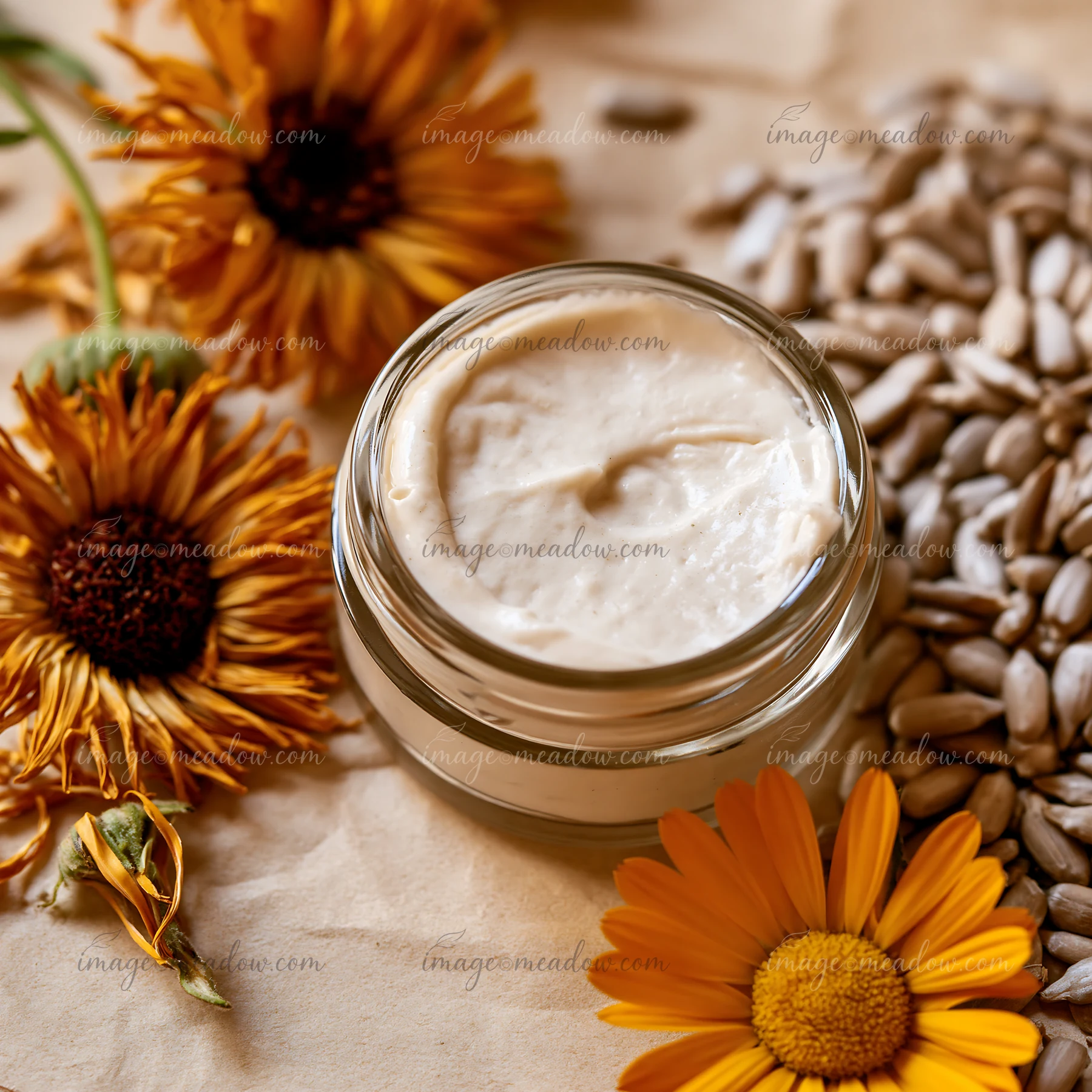 A small glass jar holds cream dose with natural skincare ingredients surrounded by sunflower seeds and marigold flowers.