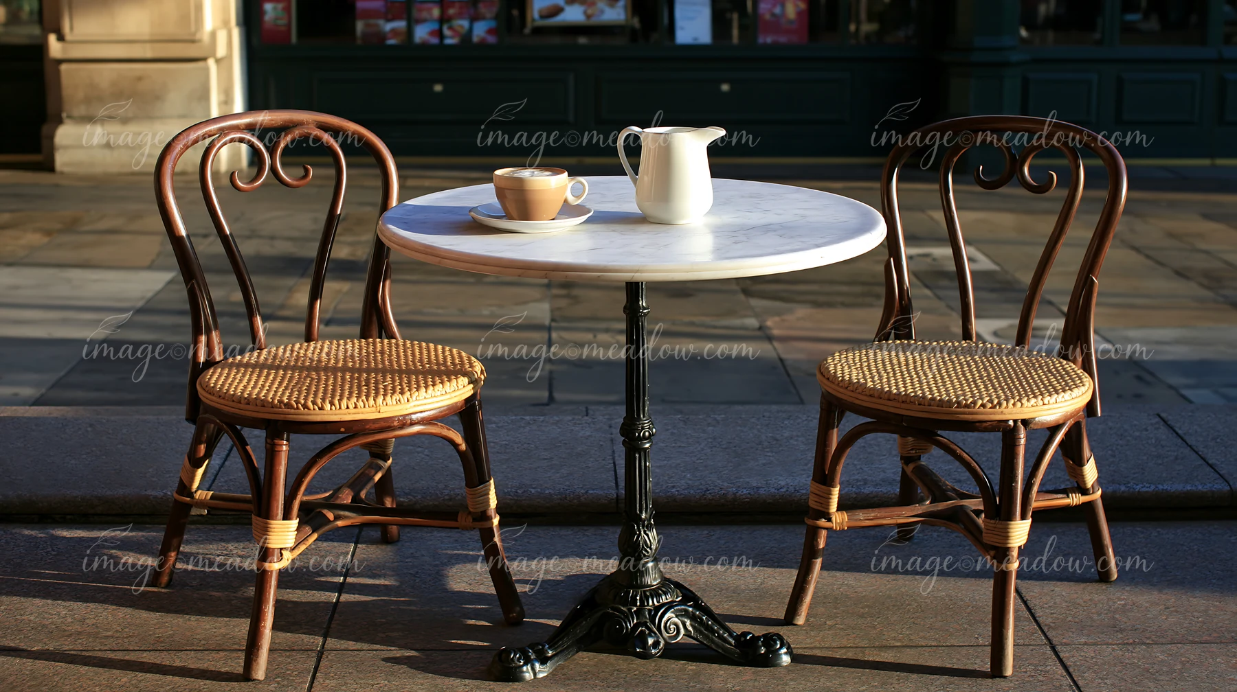 A small outdoor cafe table is set on a sidewalk with a latte and a milk jug.