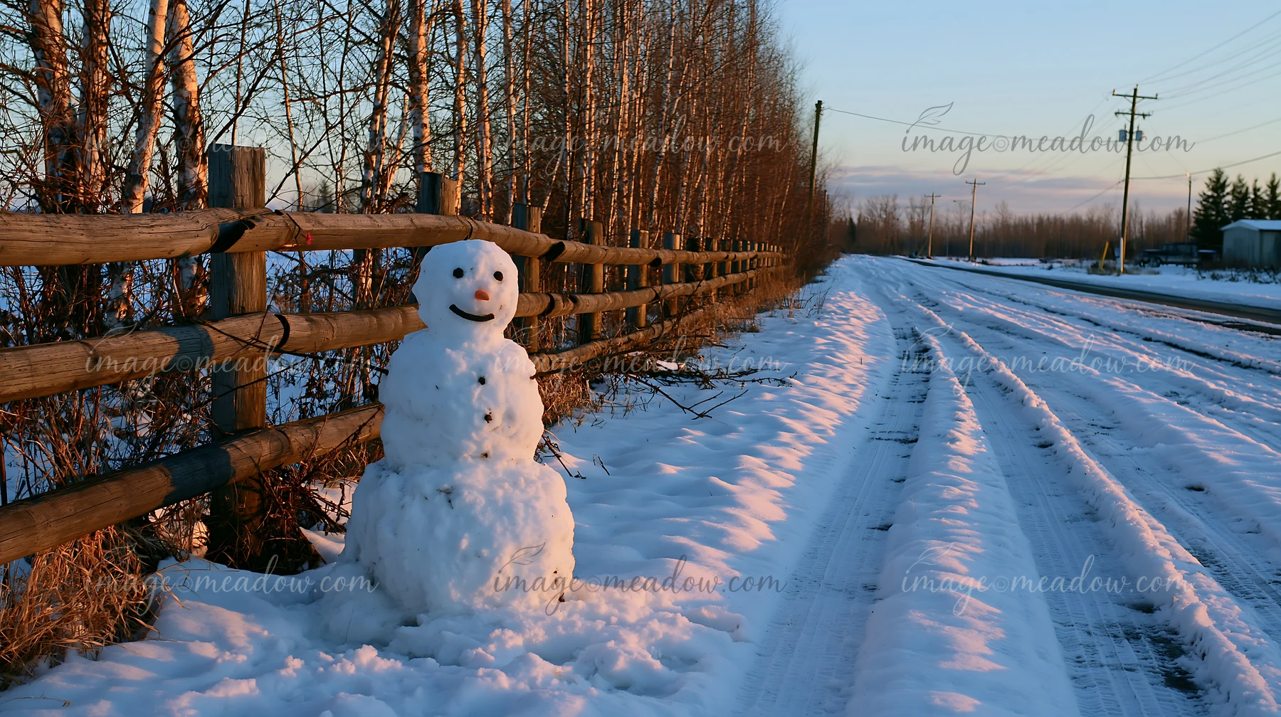 A snowman beside a wooden fence stands along a snowy road with trees in the background during sunset.