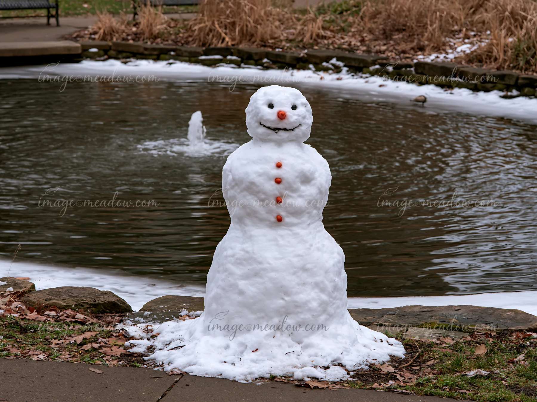 A snowman in a city park sits beside a pond with a fountain.