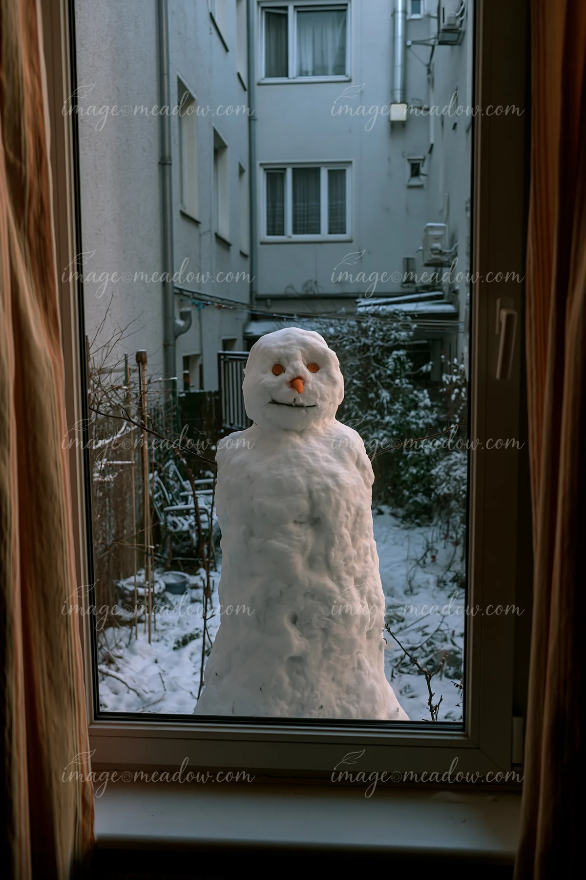 A snowman seen through a window stands in a snowy courtyard surrounded by buildings.