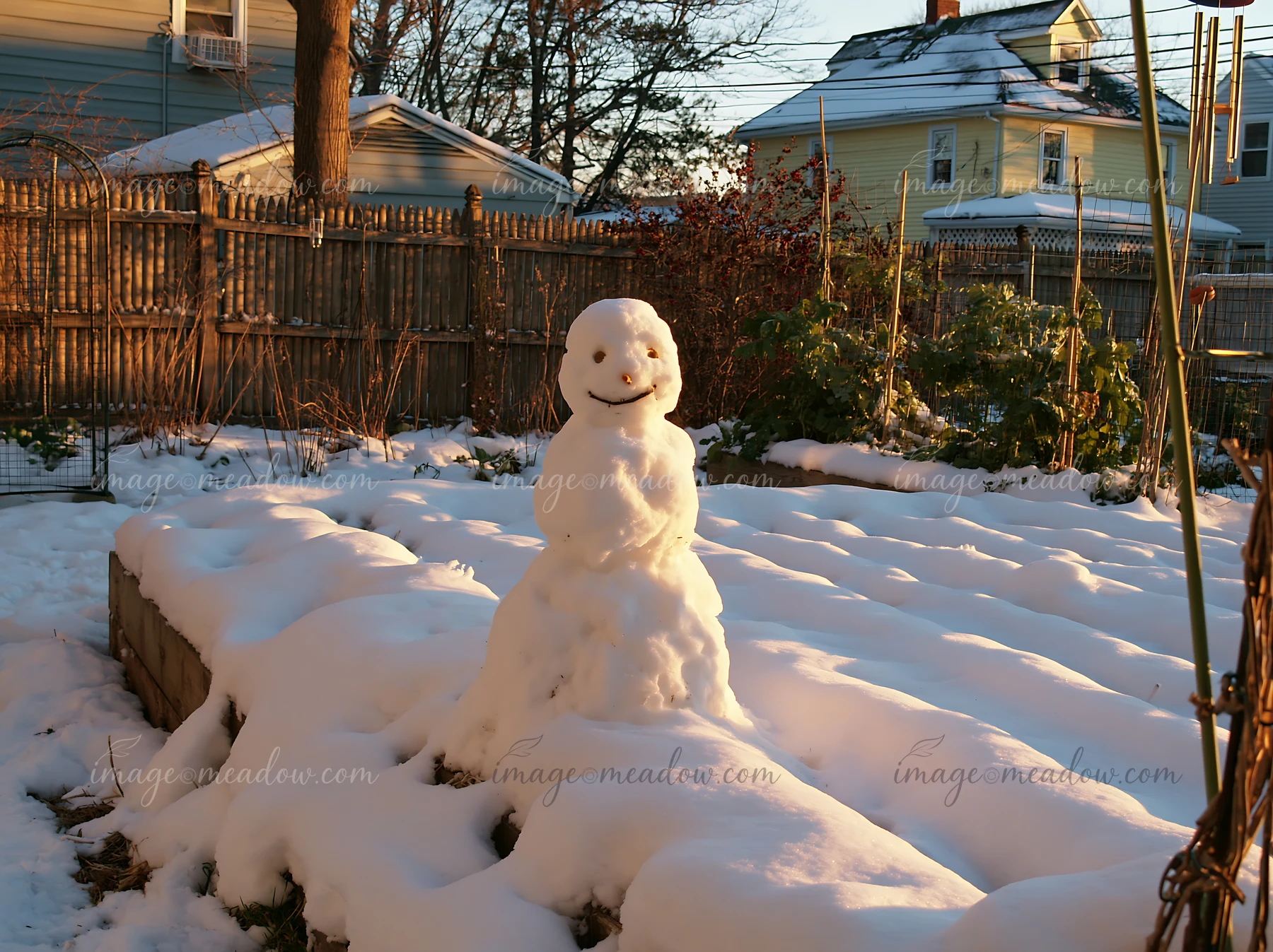 A snowman stands in a snowy backyard garden, surrounded by white snow and green plants.