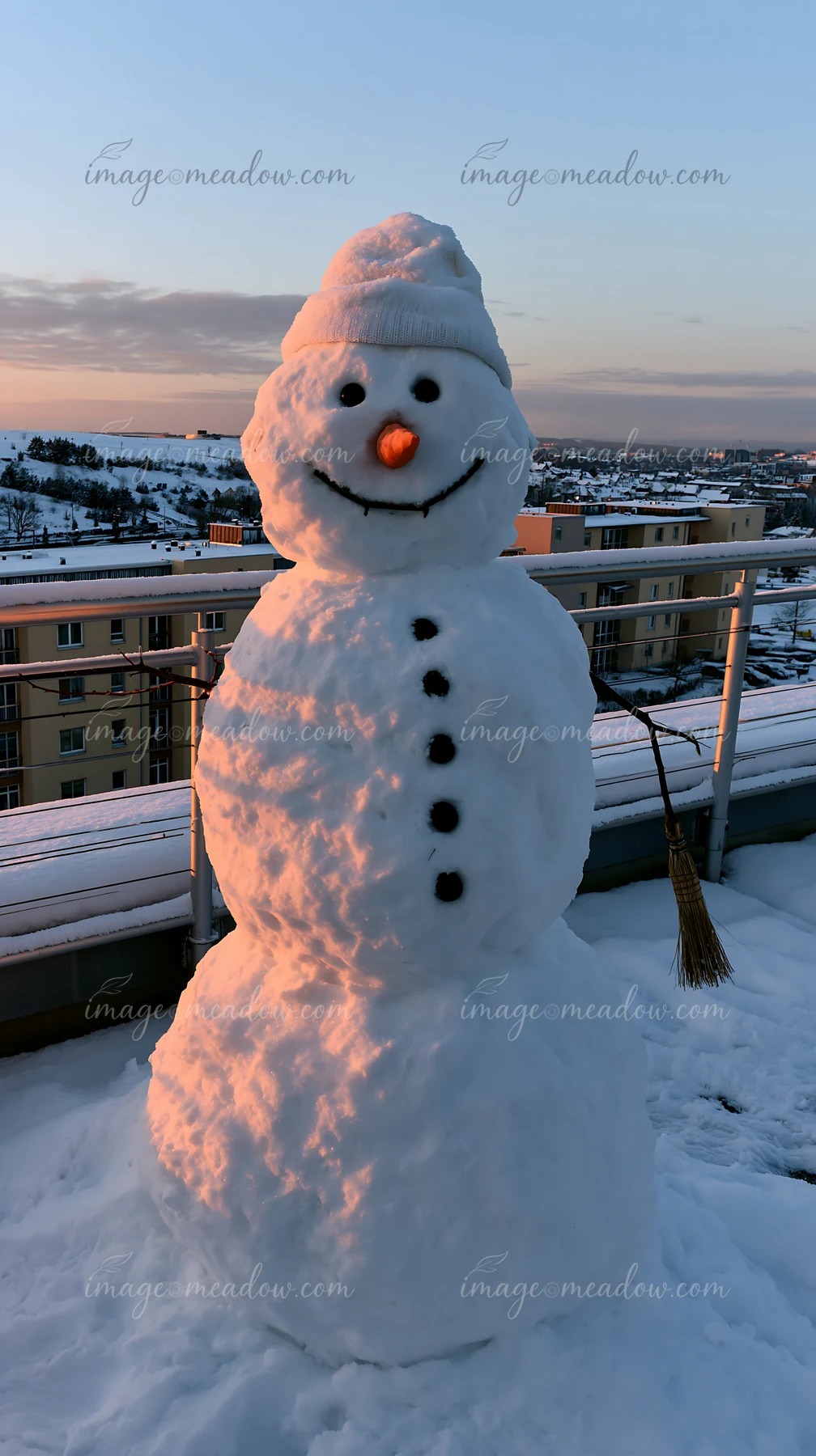 A snowman with a broomstick arm stands on a rooftop, made from snow and decorated with a carrot nose and stones.