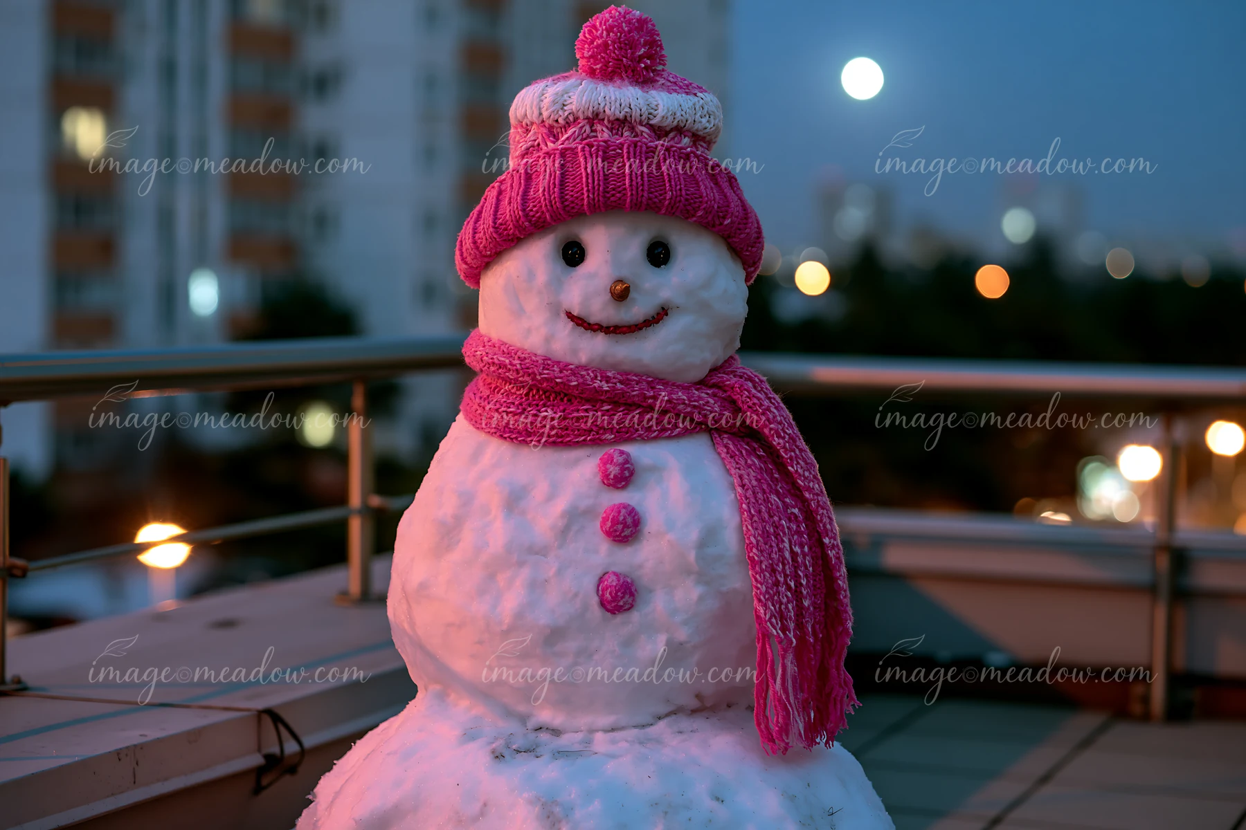 A snowman with a pink hat and scarf stands on a balcony at night under moonlight.