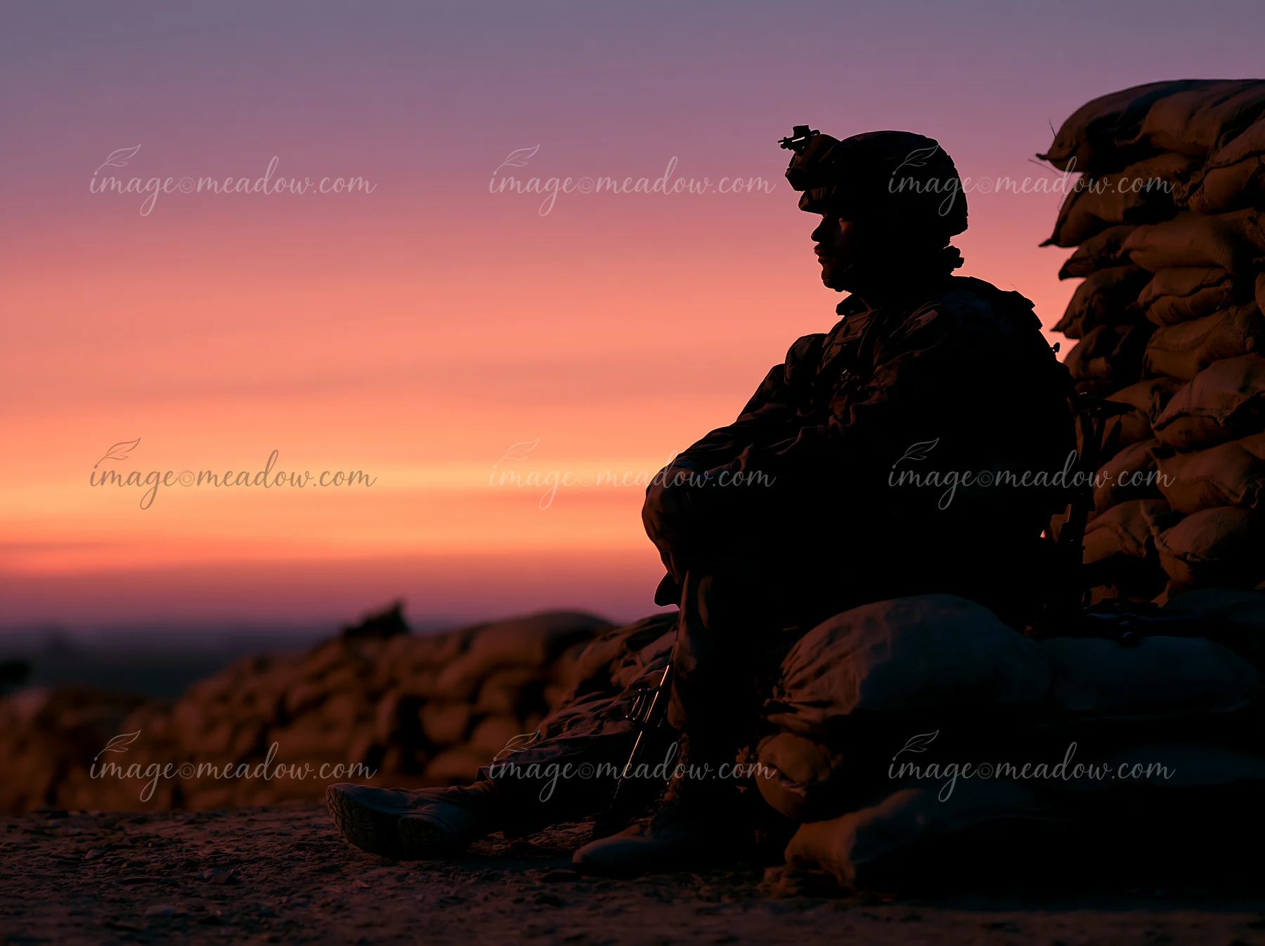 A soldier silhouette at dawn sits on sandbags, observing the horizon as the sky transitions to soft colors.