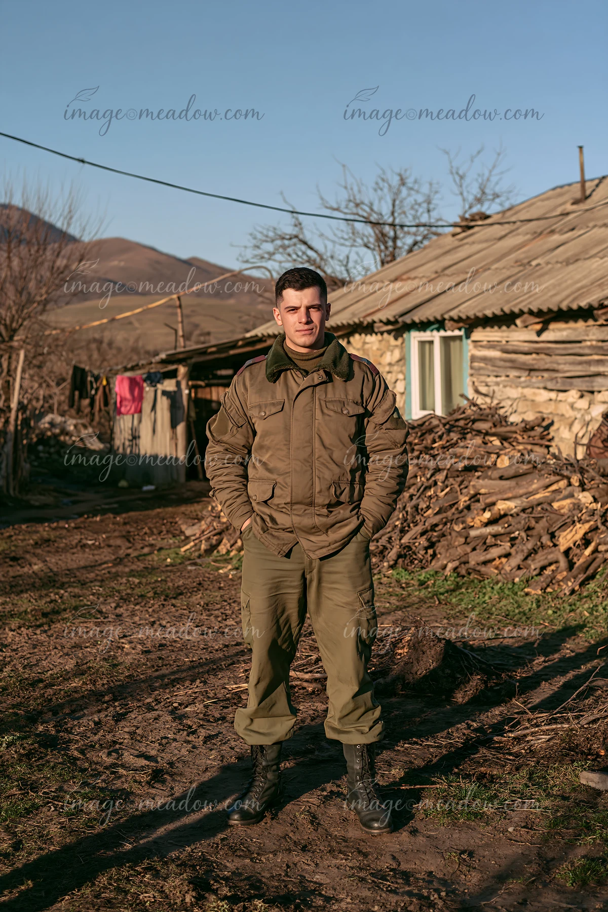 A soldier stands in front of a rustic house with a shadow on the ground.