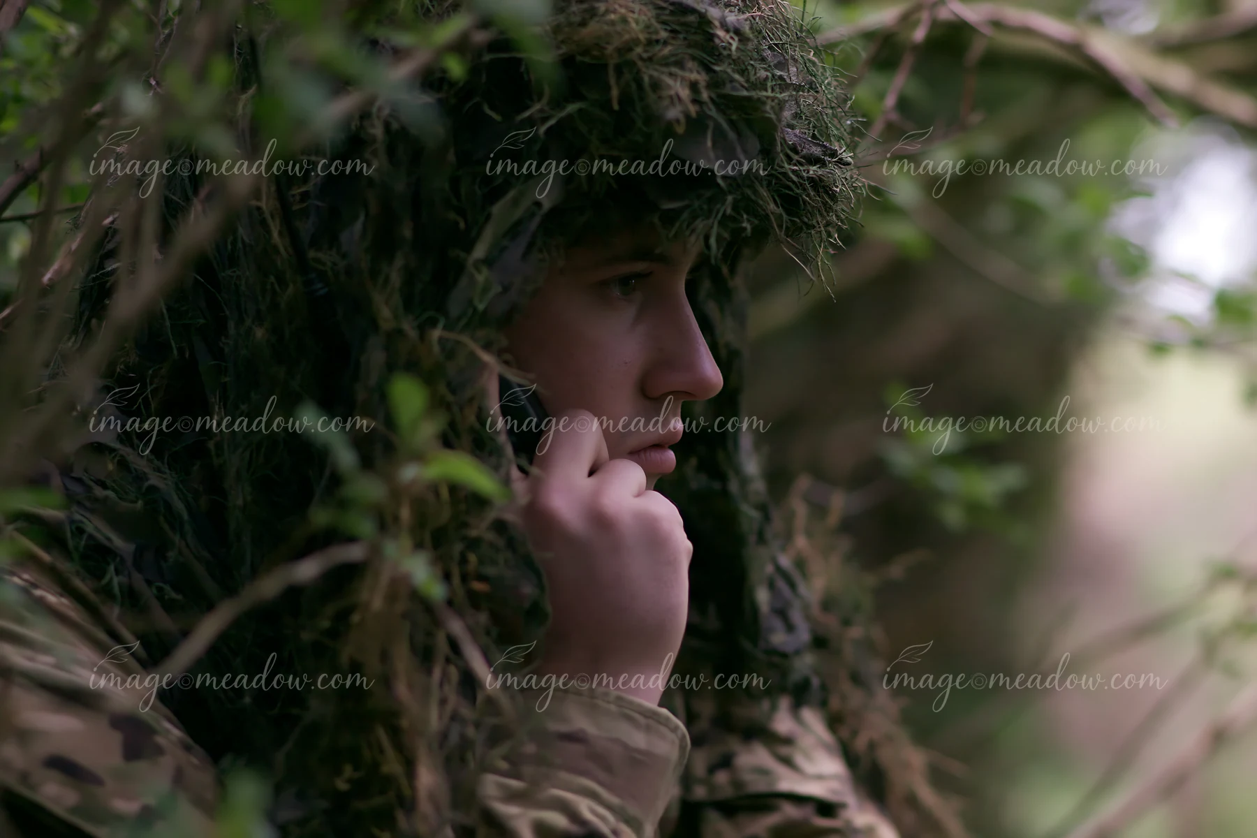 A soldier under camouflage netting speaks on the radio while hidden in dense greenery.