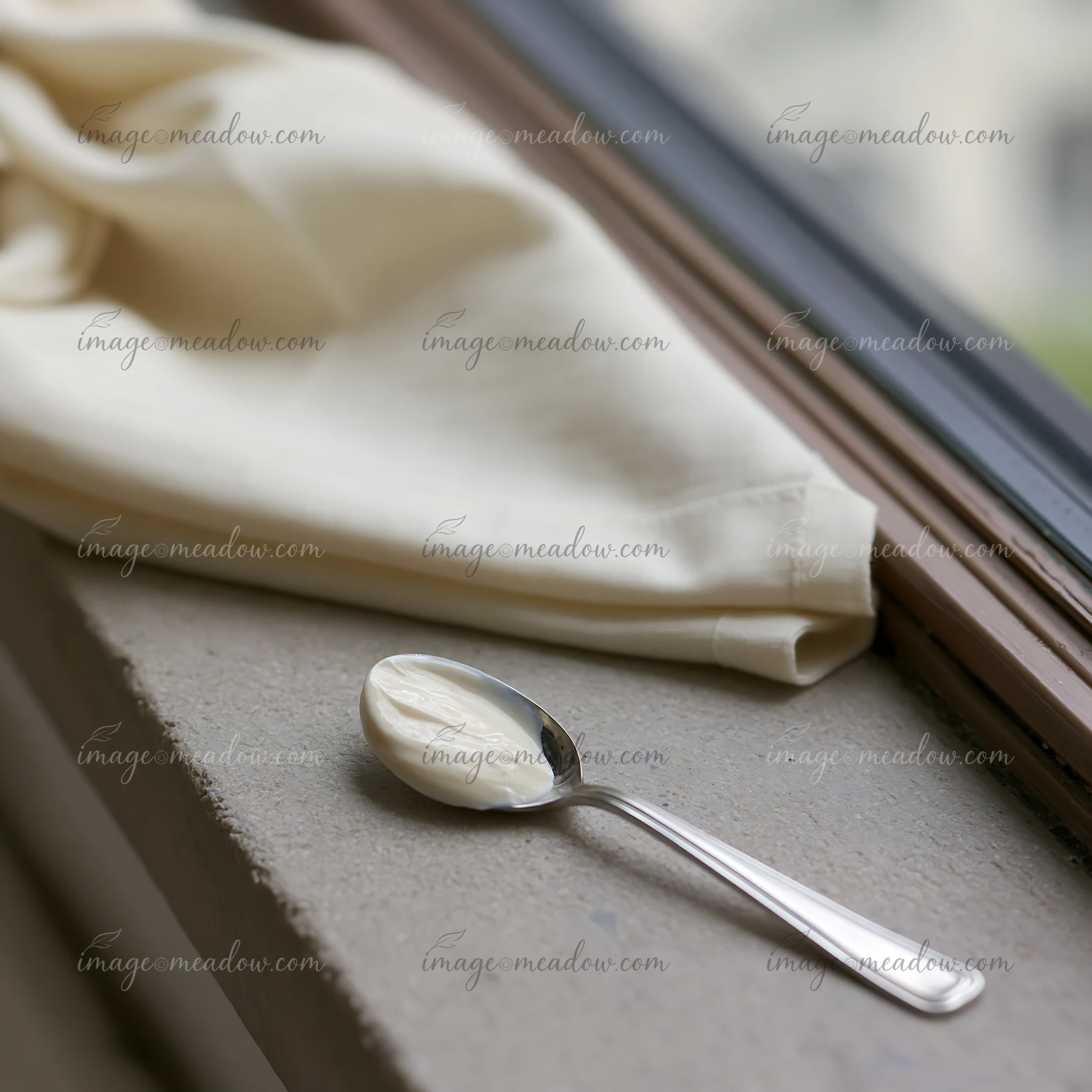 A spoon holds a small amount of cream next to a folded piece of cream-colored fabric resting on a windowsill.