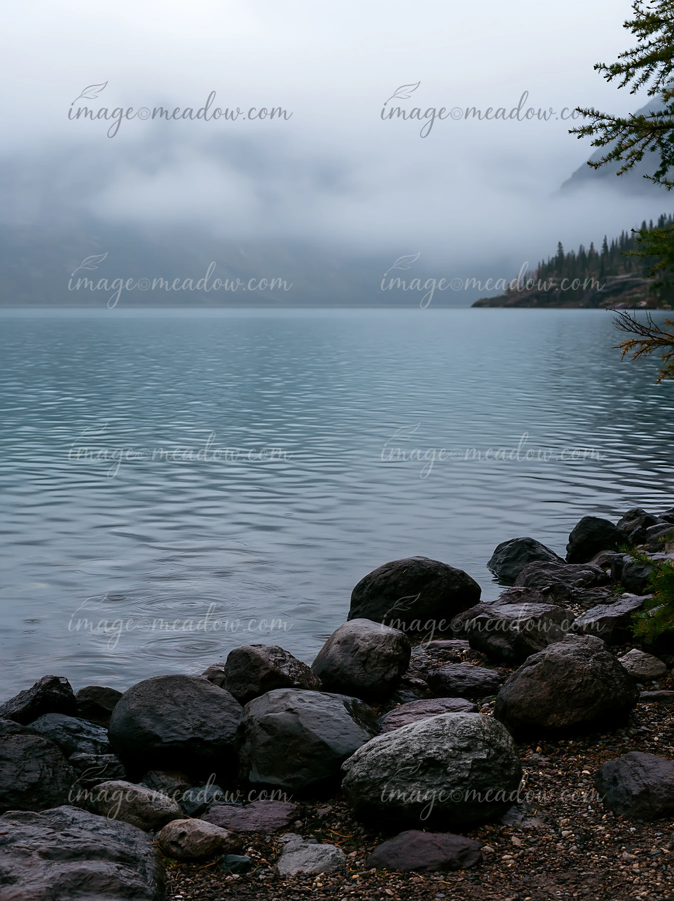 A still alpine lake sits beneath cloud-covered peaks, with dark rocks lining the shore.