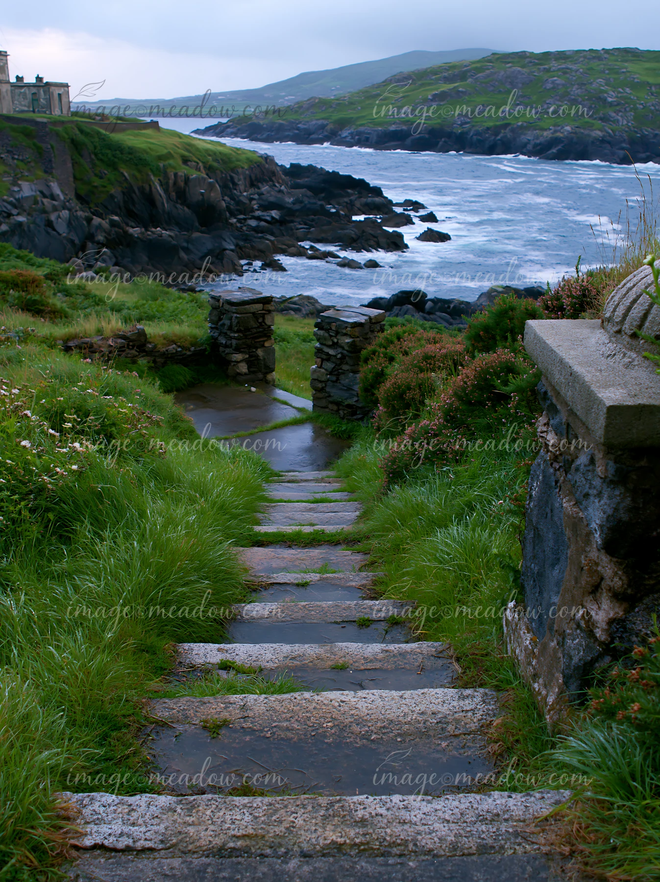 A stone pathway leads down to a rocky shoreline and an abandoned lighthouse on a remote coast.