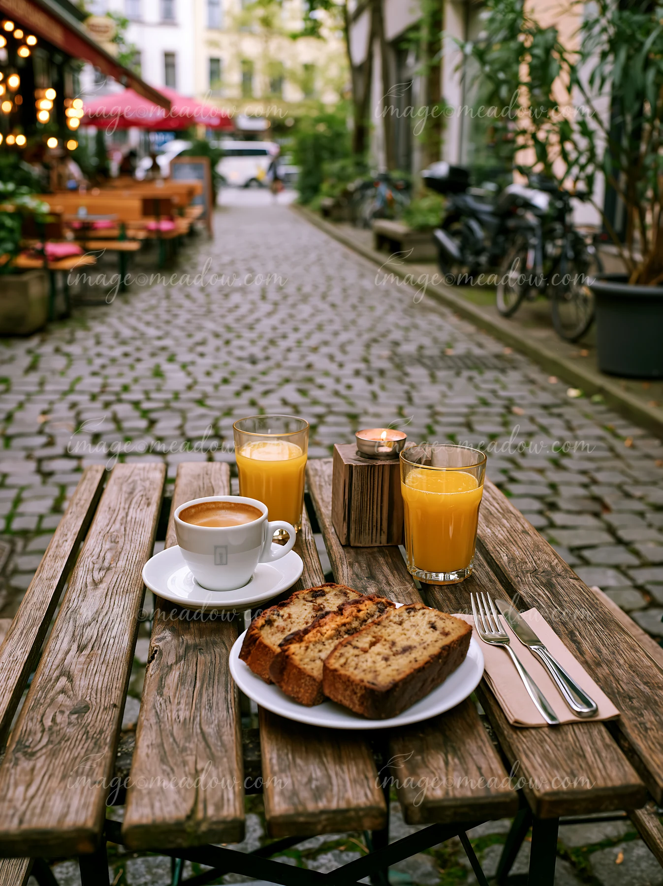 A street cafe table is set with a cup of milk coffee, slices of bread, and glasses of juice.