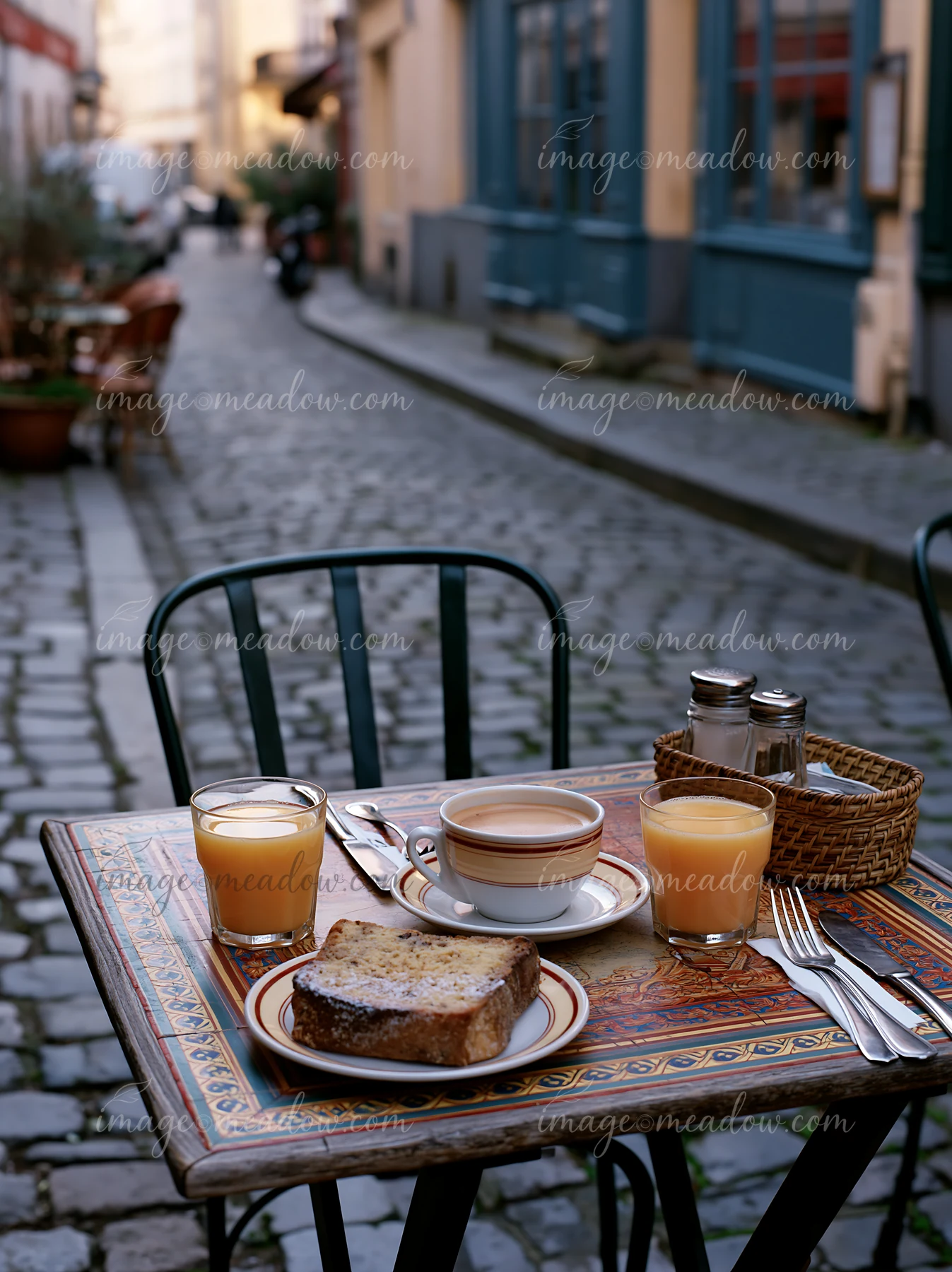 A street cafe table is set with milk coffee, fresh juice, and breakfast bread.