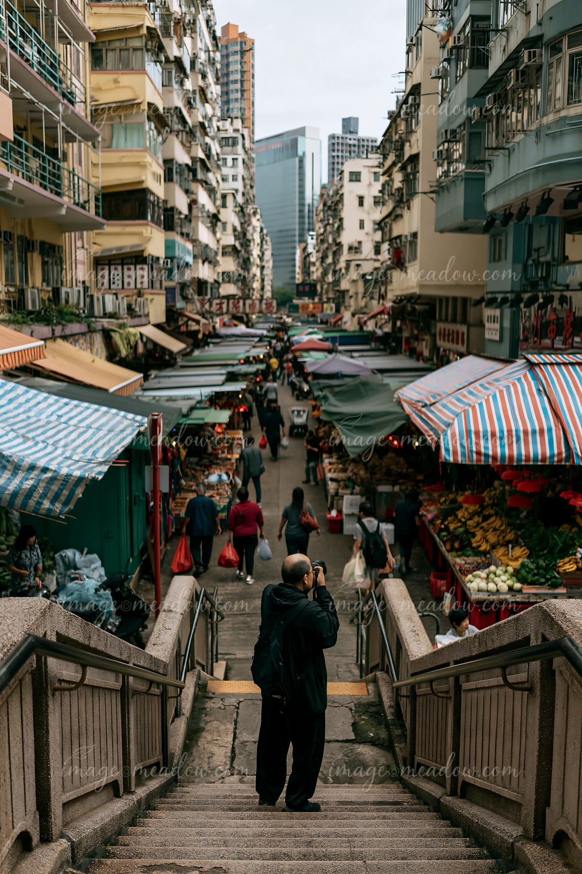 A street photographer taking pictures in an urban market stands at the top of stairs overlooking stalls filled with fresh produce and shoppers.