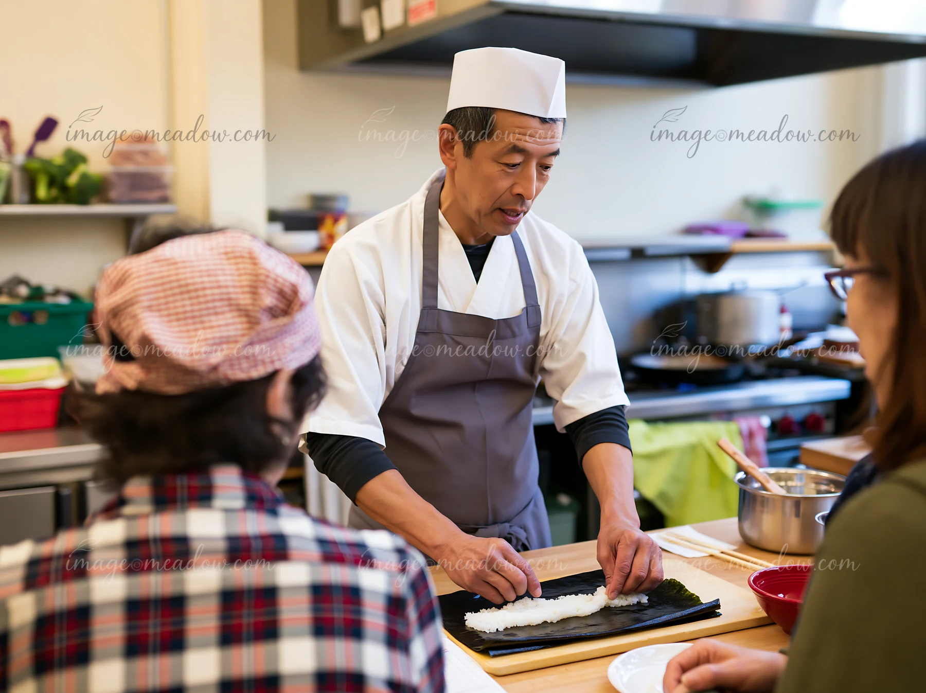 A sushi chef demonstrates traditional sushi preparation techniques to adults in a kitchen setting.