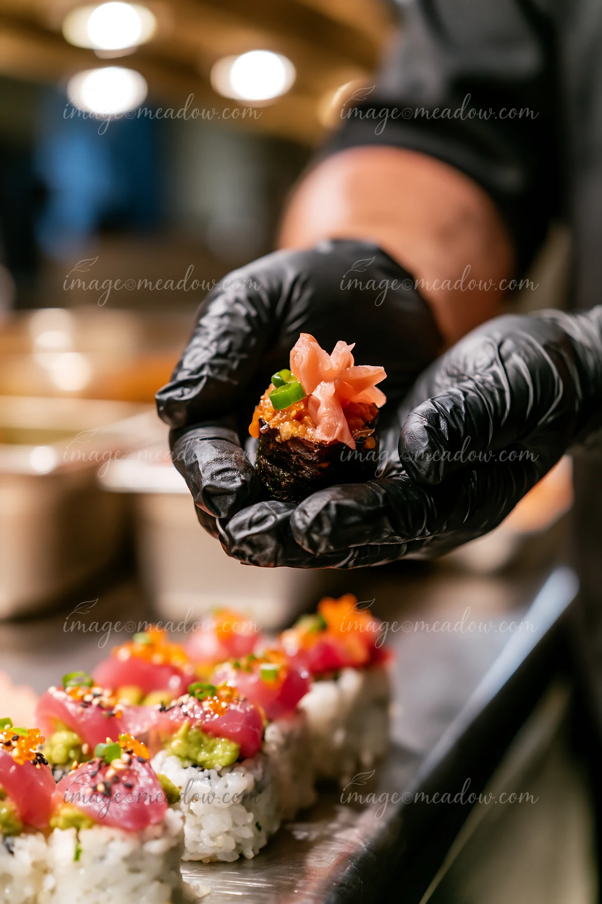 A sushi chef presents one sushi piece with pickled ginger while preparing sushi in a restaurant kitchen.