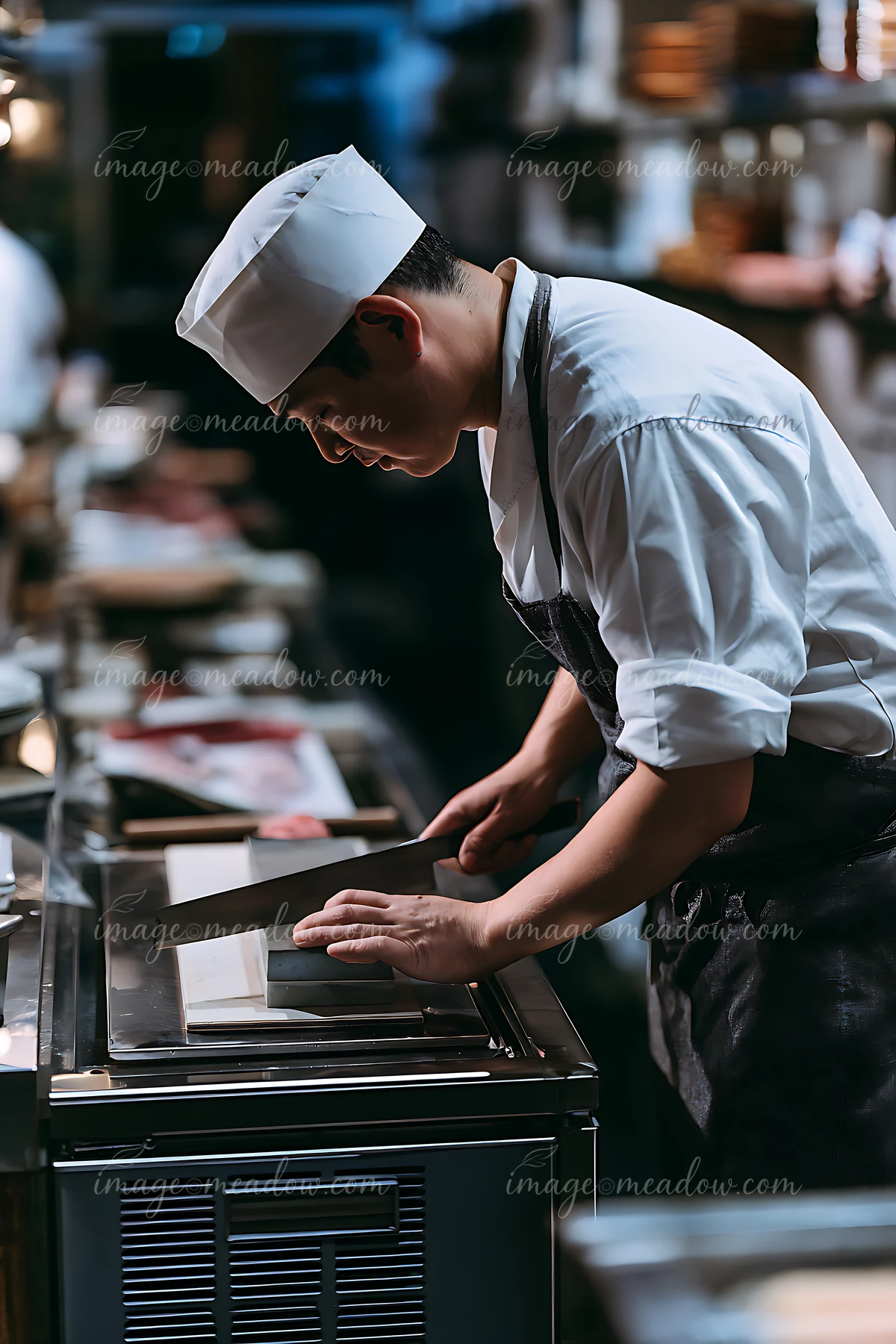 A sushi chef sharpens a knife on a countertop in a busy professional kitchen.