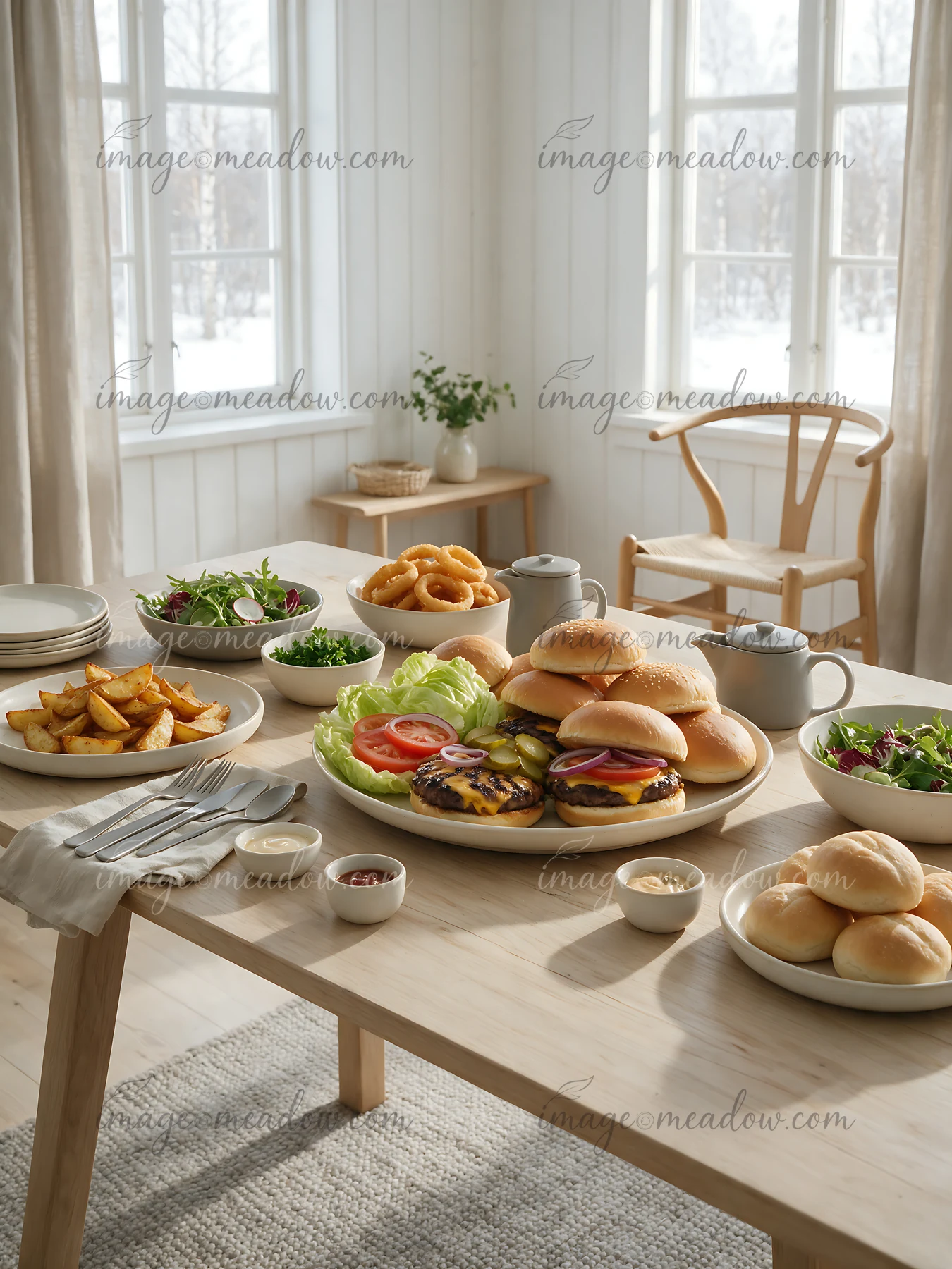 A table displays a burger buffet setup with burgers, fries, salads, and sauces in a bright room with windows.