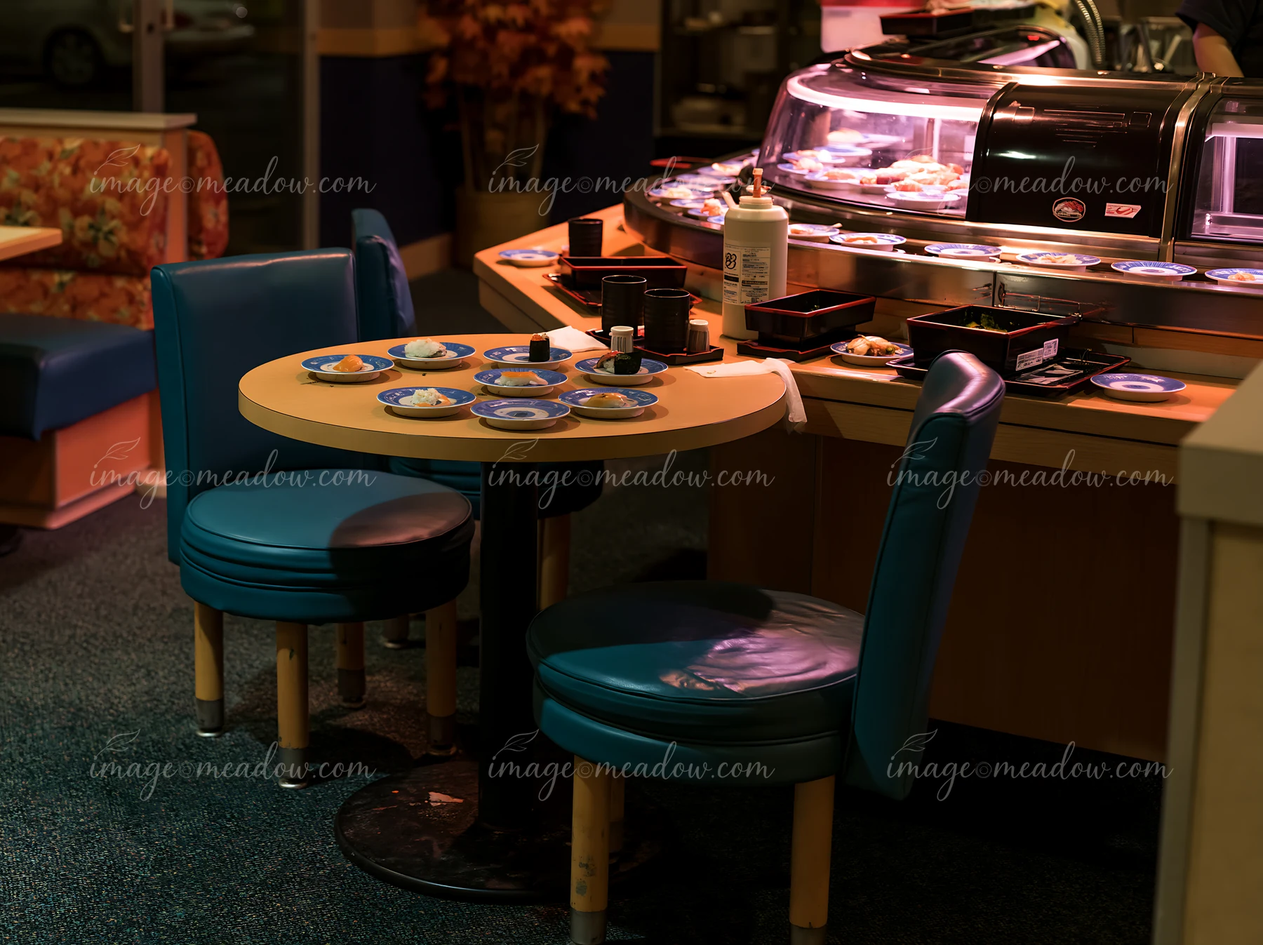 A table with plates of sushi sits near a busy sushi conveyor belt restaurant.
