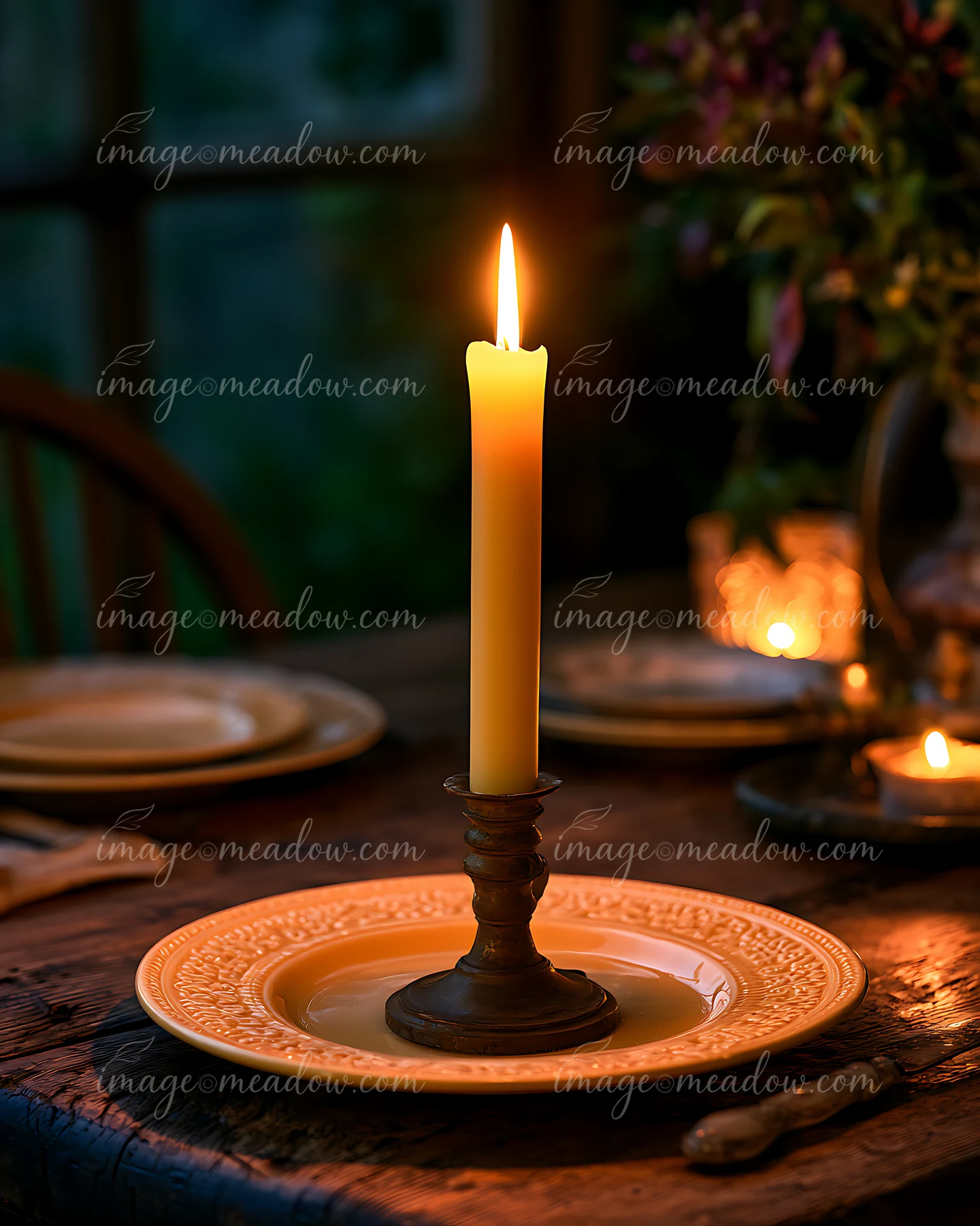 A tall candle stands in a holder on a plate, illuminating a rustic dining table.